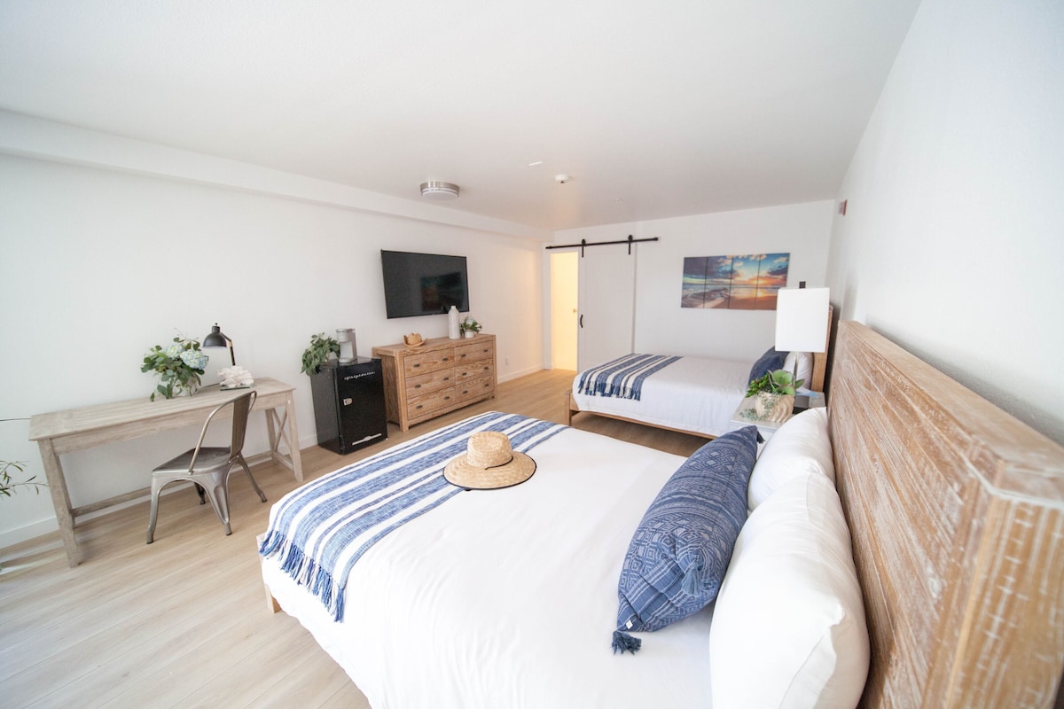 A bedroom with a desk, two beds, a TV, and a painting.