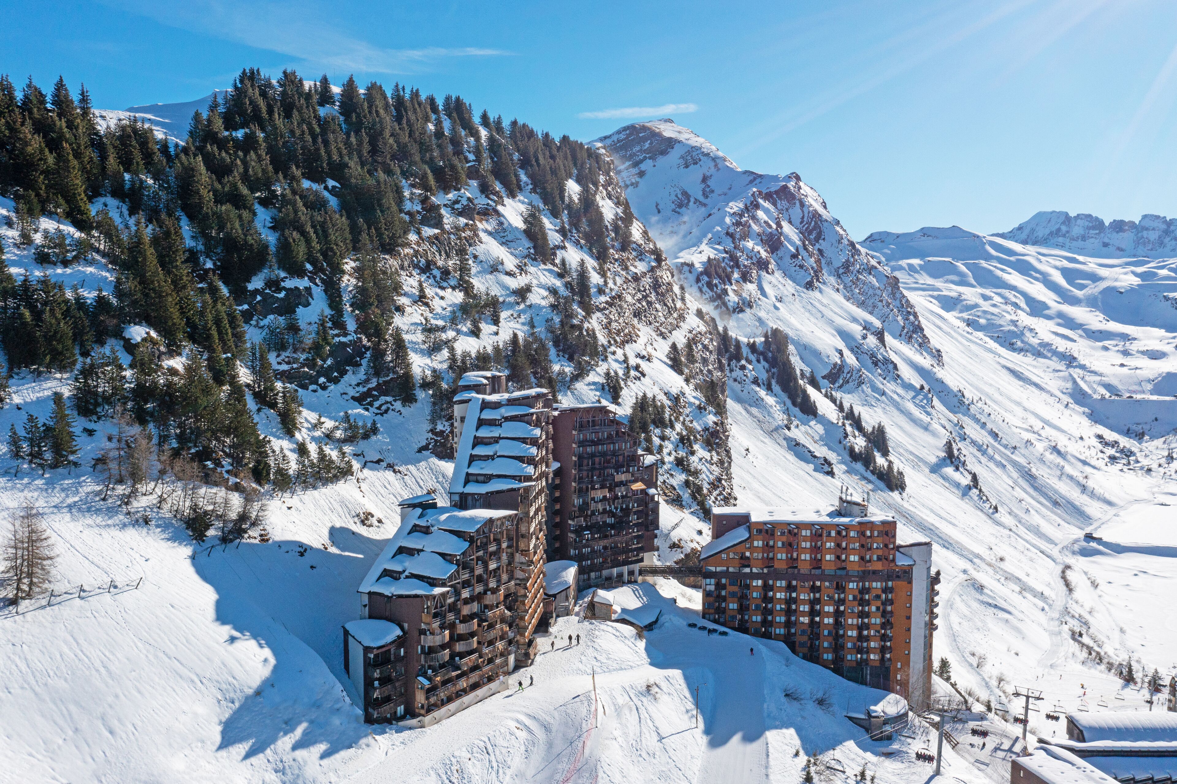 A snow-covered mountain resort with multiple buildings, including a hotel with balconies and a ski lift.