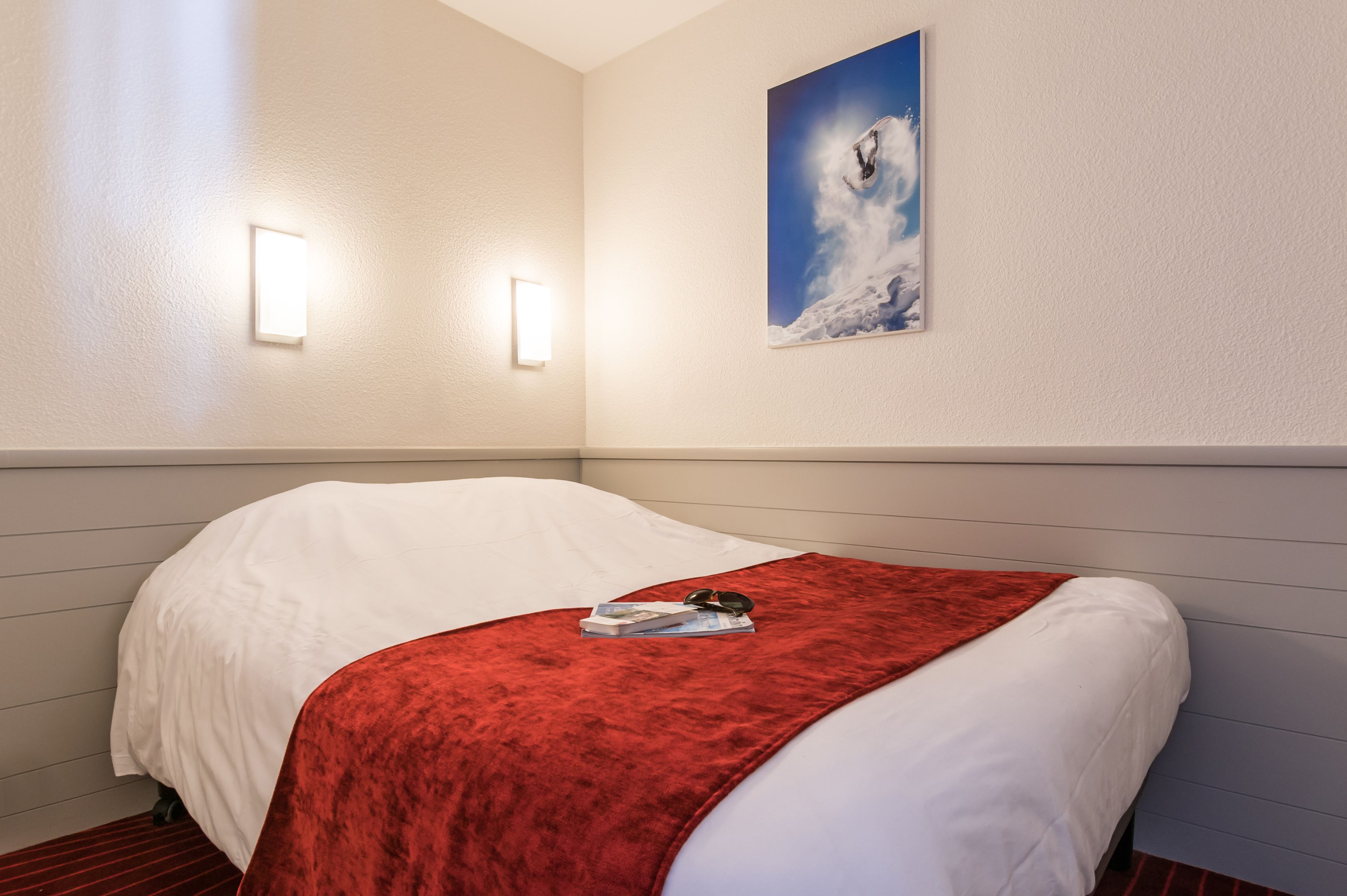 A neatly made bed with a white and red blanket, a framed picture on the wall, and two wall-mounted lights.
