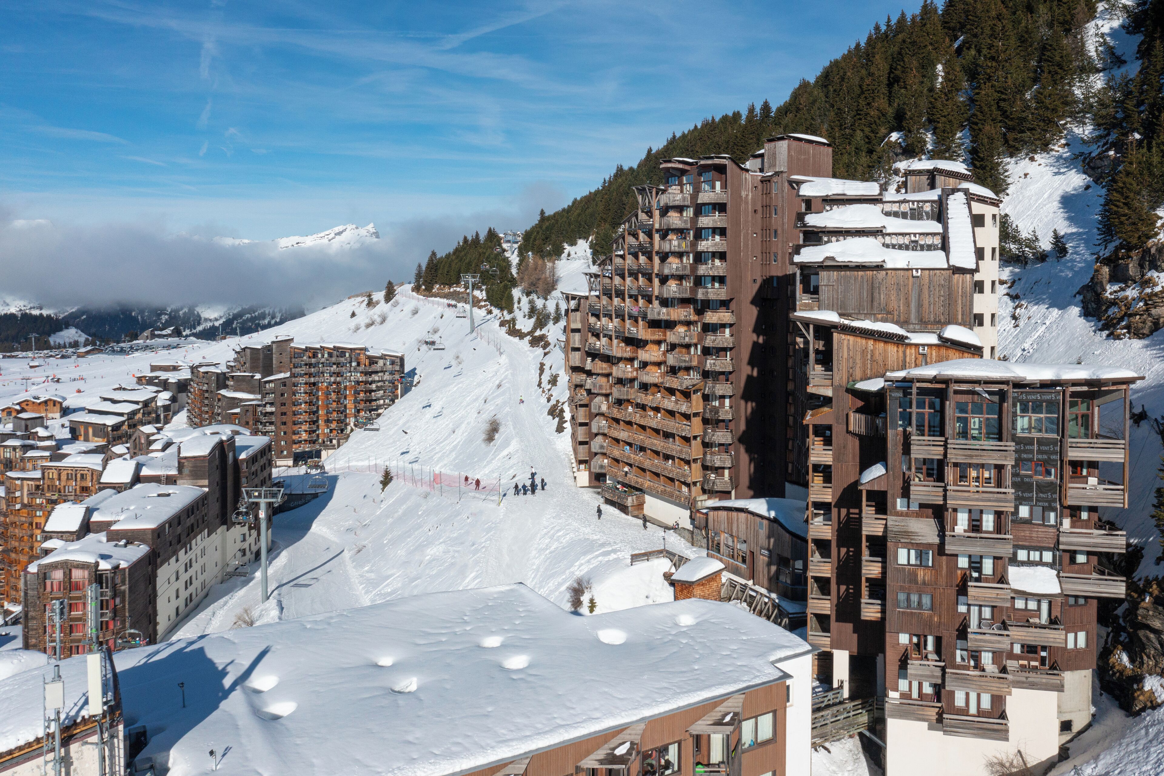A snow-covered mountain town with multi-story buildings, including a prominent hotel with balconies and ski slopes in the background.