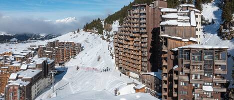 A snow-covered mountain town with multi-story buildings, including a prominent hotel with balconies and ski slopes in the background.