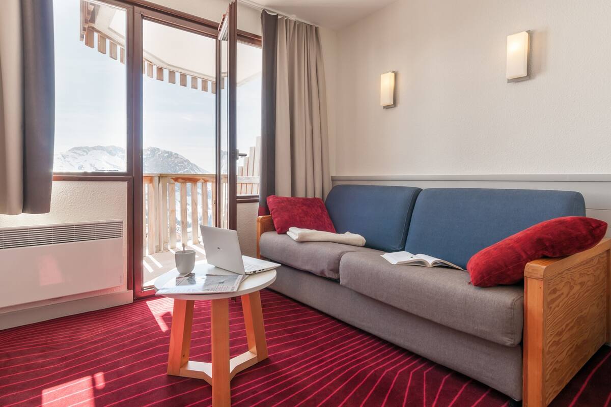 Ein Hotelzimmer mit Balkon, einer Couch mit roten Kissen, einem kleinen Tisch mit einem Laptop und einem roten Teppichboden.