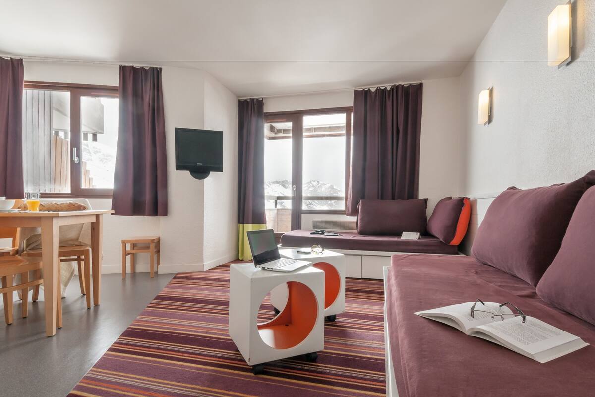 Ein modernes Hotelzimmer mit Essbereich, einer Couch, einem kleinen Tisch mit einem Laptop und einem großen Fenster mit Vorhängen.
