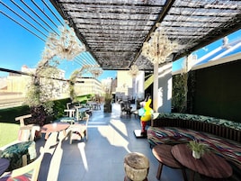 Un patio al aire libre con varios conjuntos de mobiliario, incluyendo mesas y sillas, bajo una pérgola sombreada.