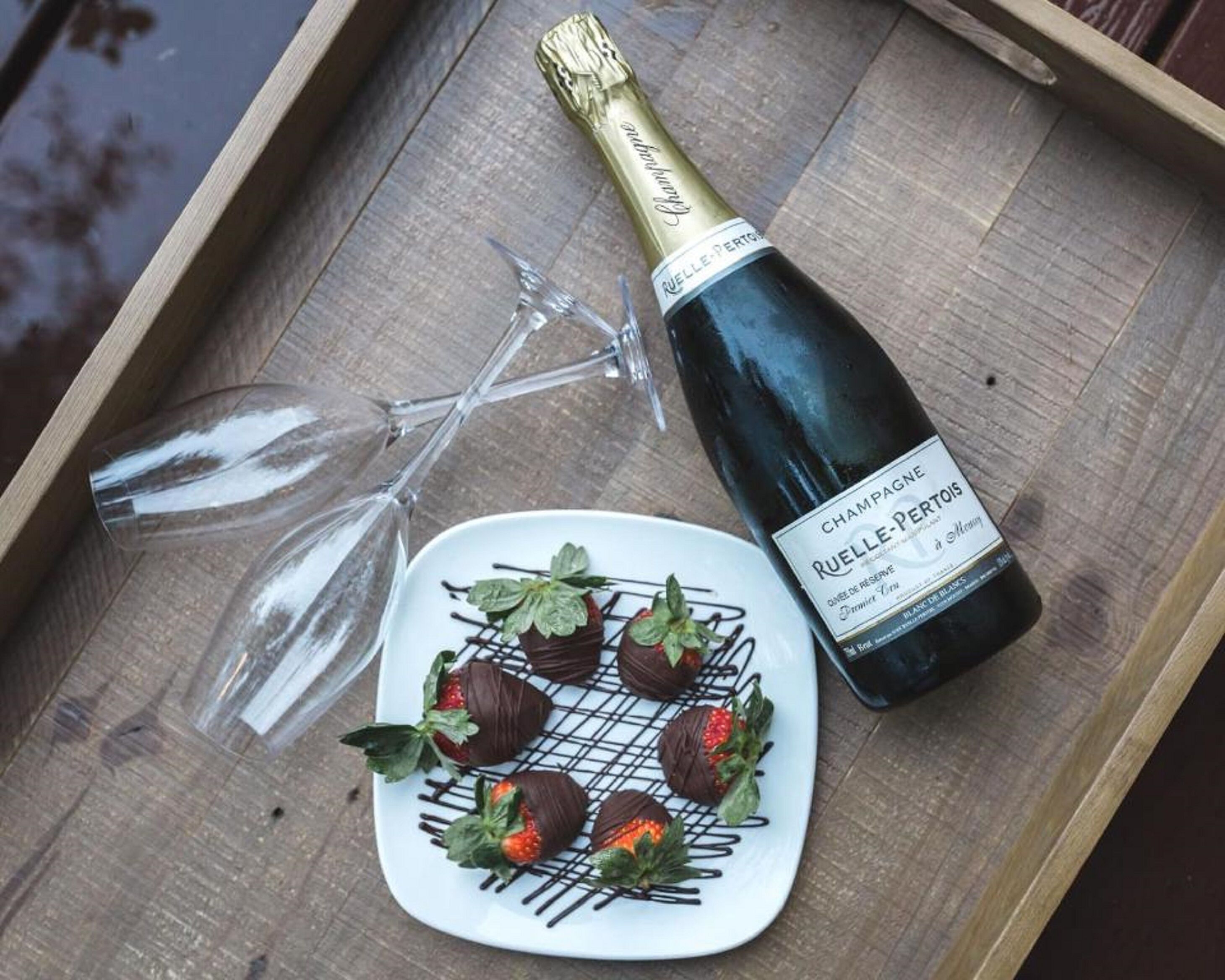 Champagnefles, een bord met chocoladebedekte aardbeien en twee lege wijnglazen op een houten dienblad.