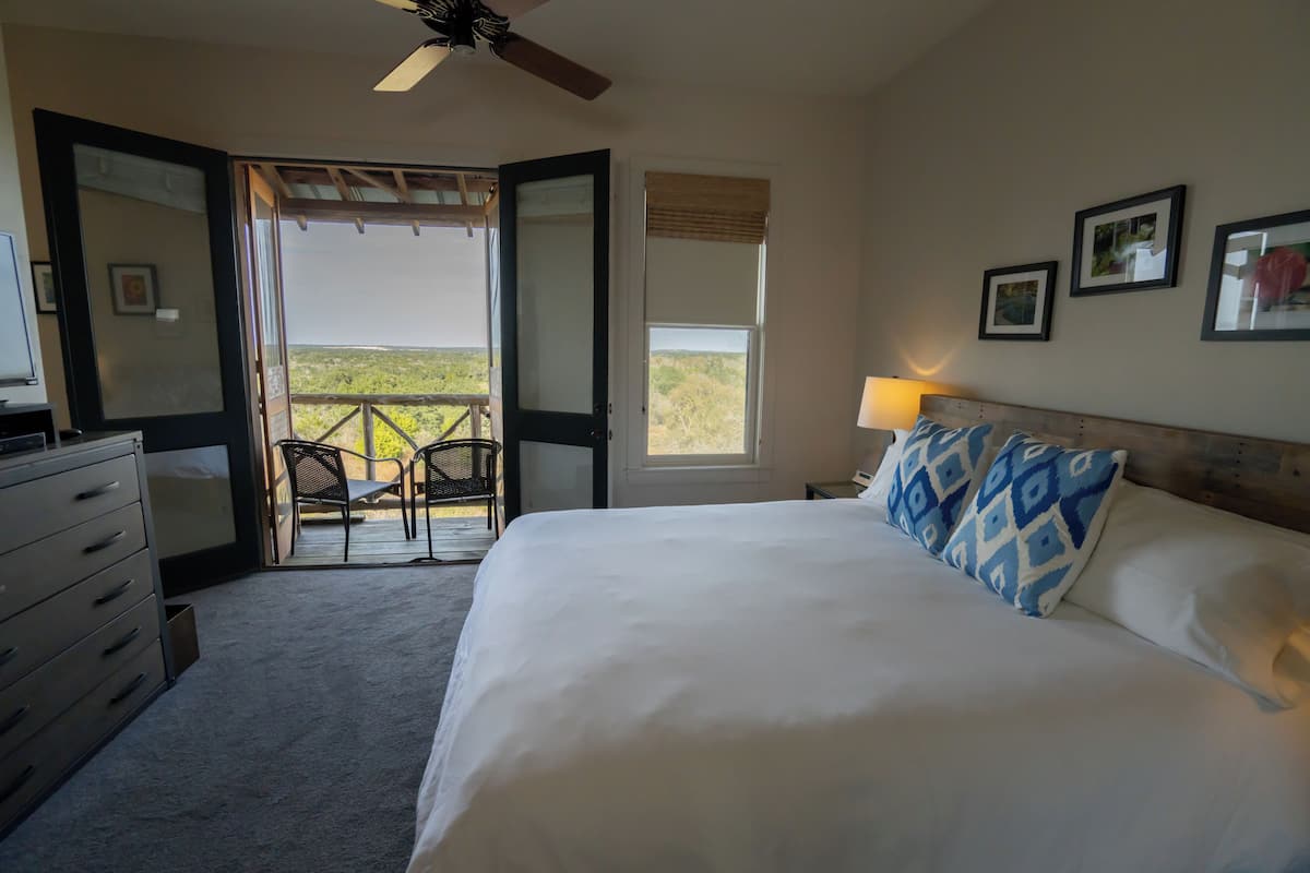 A bedroom with a large bed, a dresser, a ceiling fan, a window with a view, and a balcony with chairs.