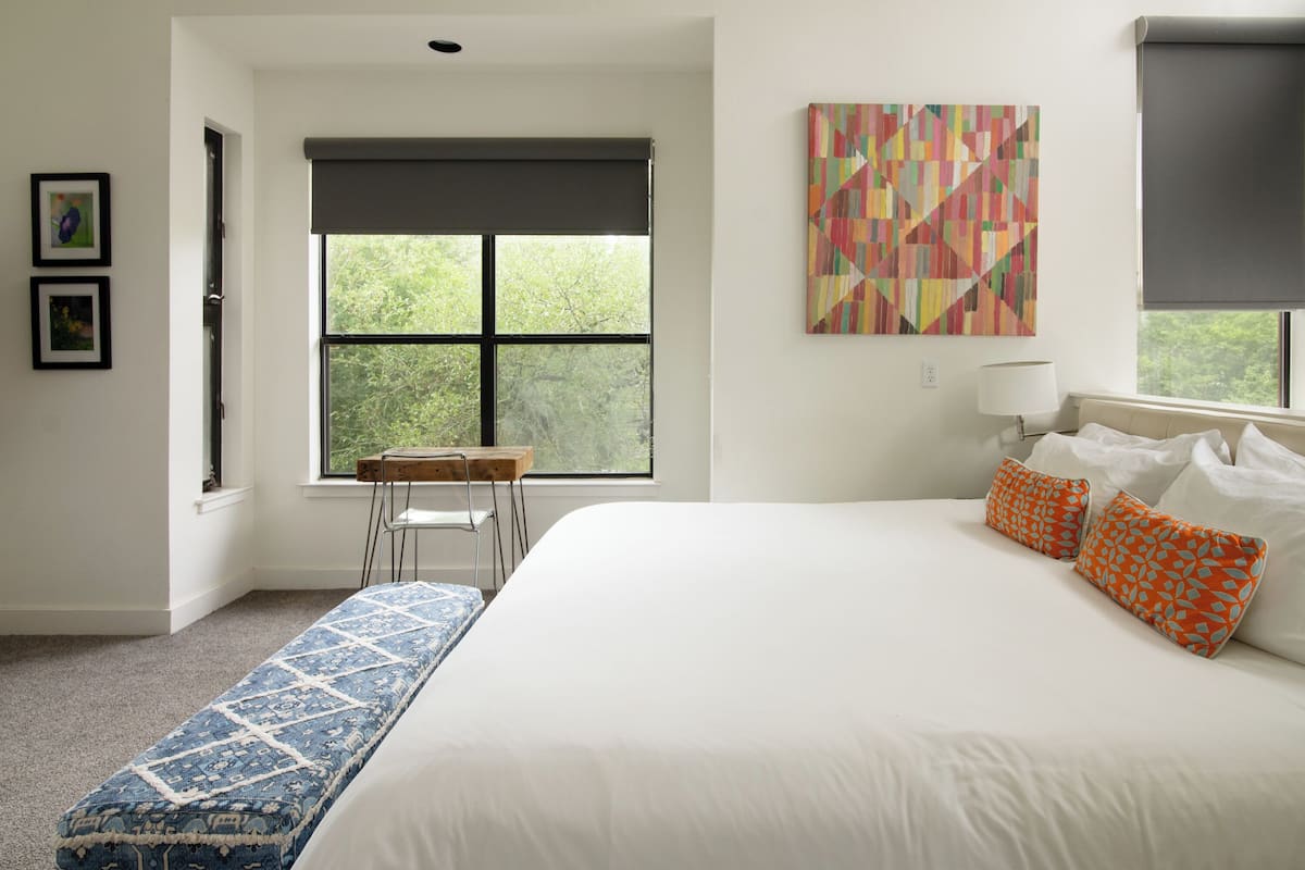 A bedroom with a large bed, two pillows, a bench, a chair, and a window with blinds.