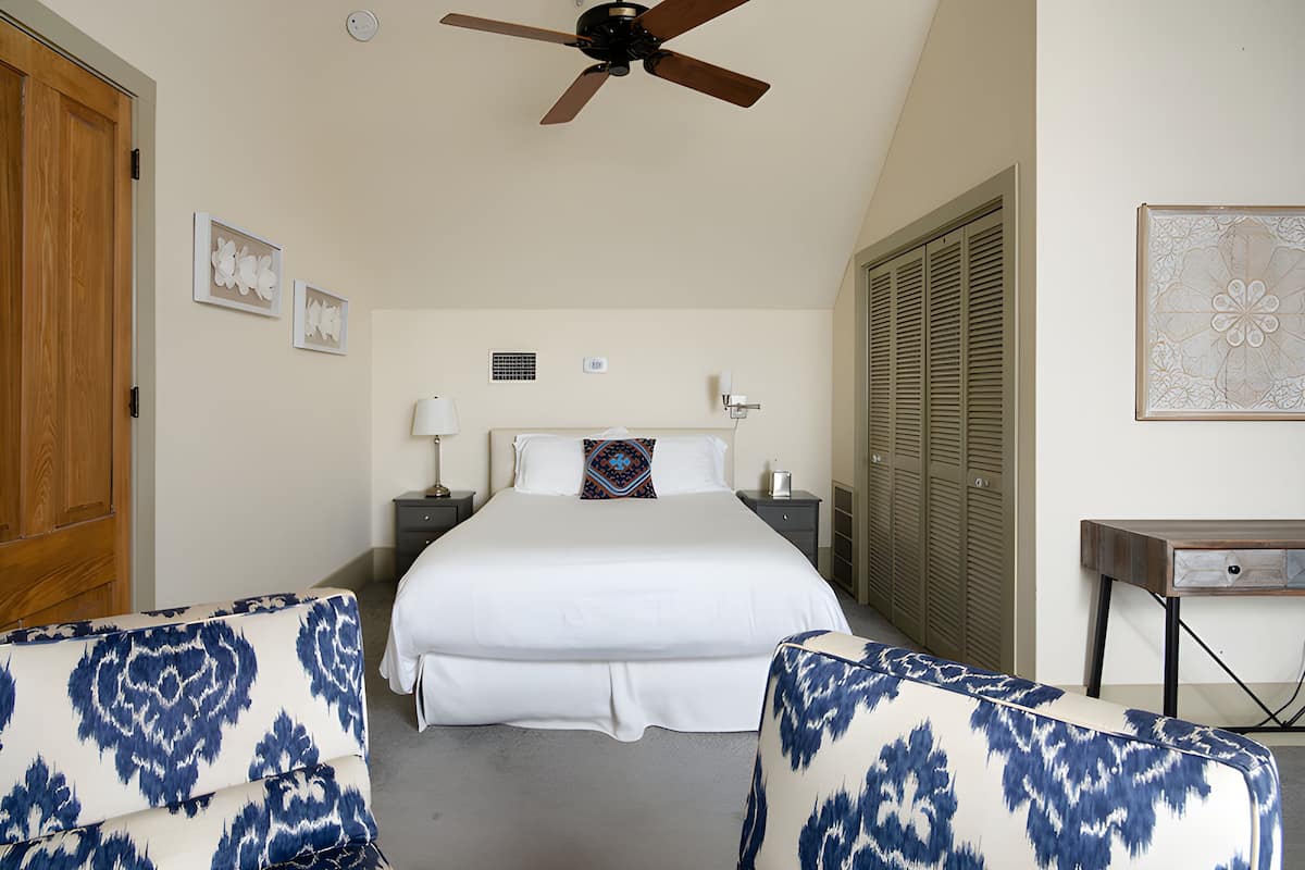 A bedroom with a large bed, two nightstands, a ceiling fan, a wooden door, and a small table with a lamp.