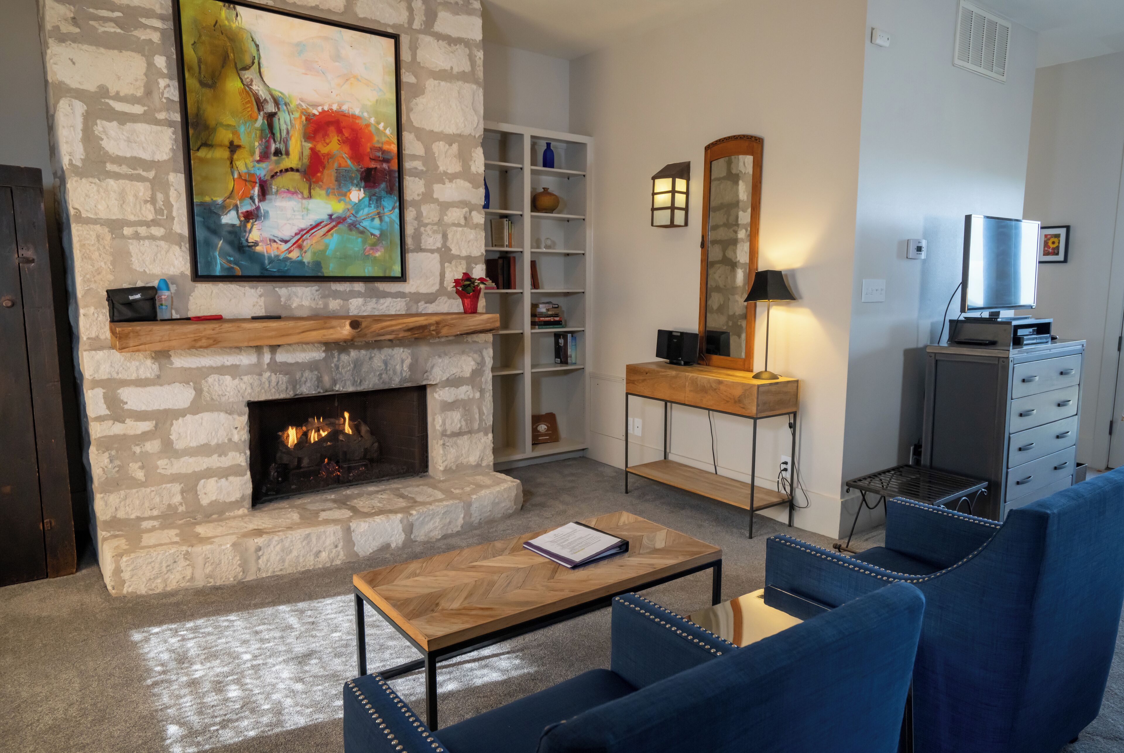 A living room with a stone fireplace, a bookshelf, a television, and blue upholstered chairs.