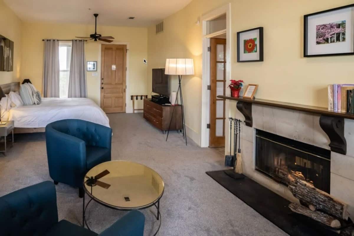 A hotel room with a bed, a blue armchair, a small round table, a fireplace, and a bookshelf.