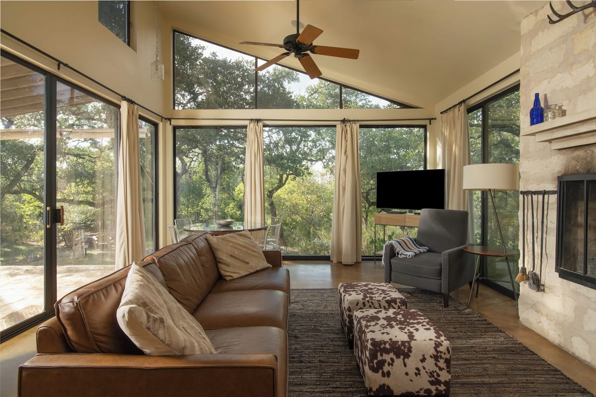 A spacious living room with a leather sofa, a patterned ottoman, a grey armchair, and a flat-screen TV.