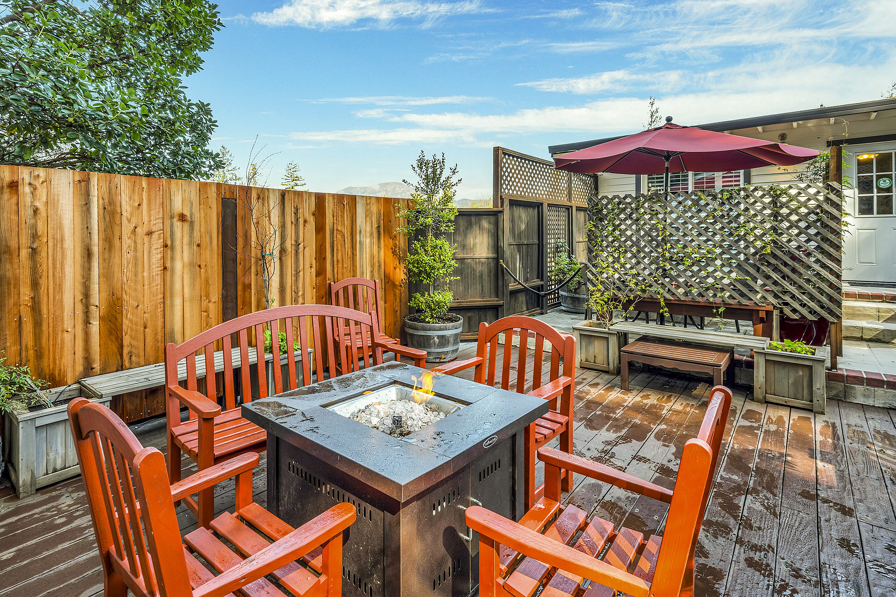 A wooden deck with a fire pit table surrounded by wooden chairs, a wooden fence, and a lattice structure with a red umbrella.