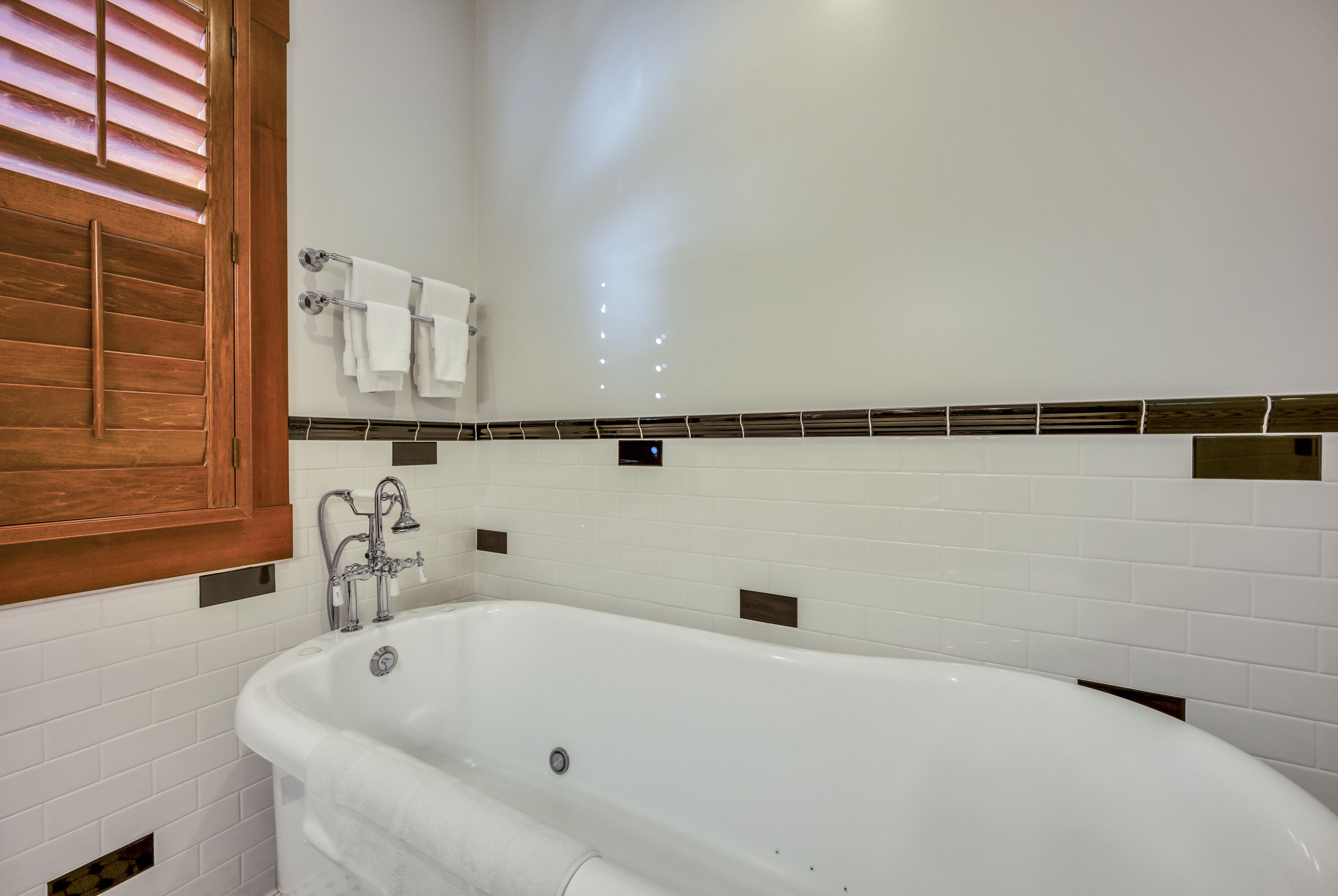 A modern bathroom with a freestanding bathtub, white tiled walls, and a wooden window shutter.
