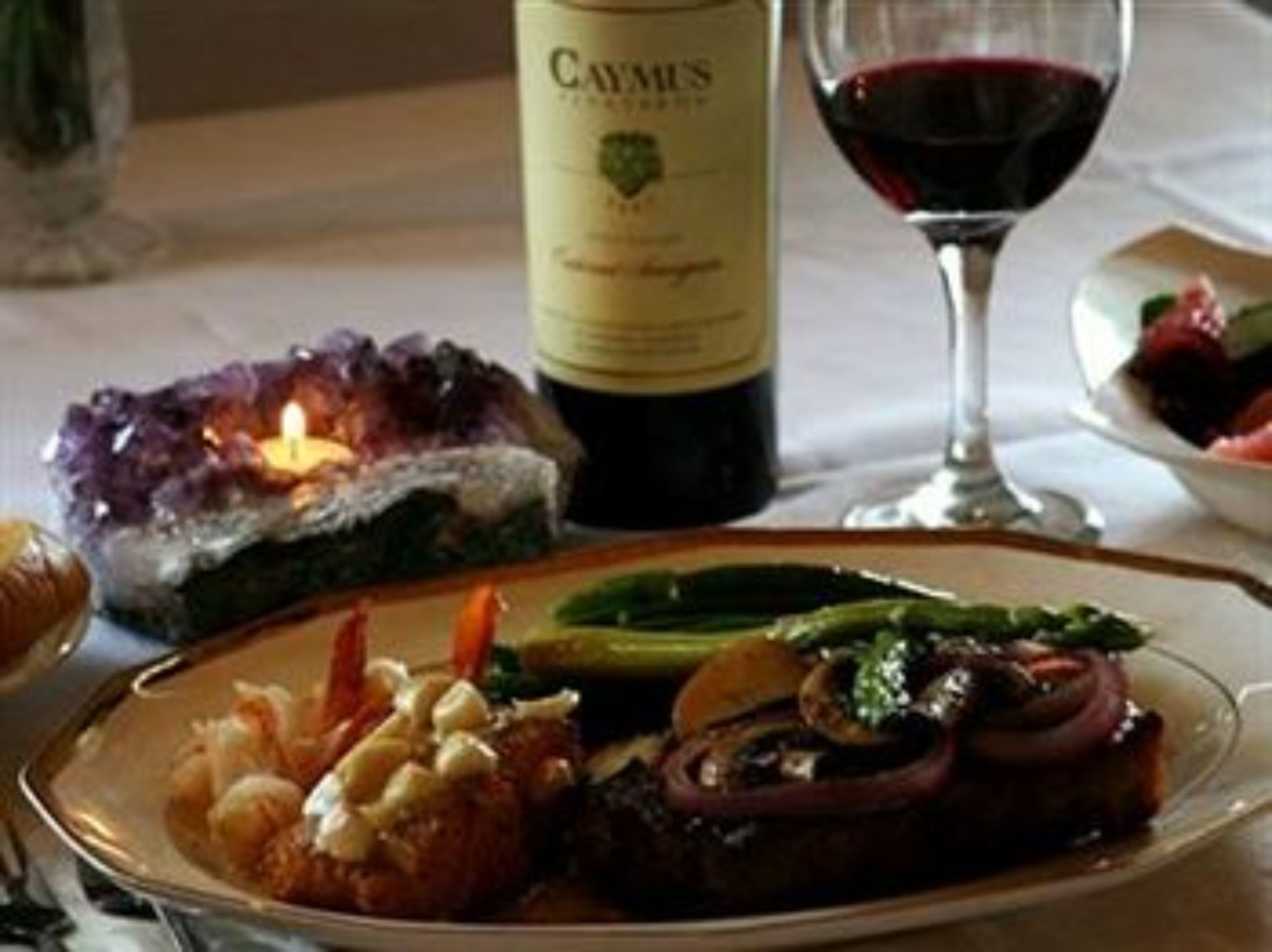 A dining table with a bottle of Caymus wine, a glass of red wine, and a plate of food including asparagus and mushrooms.