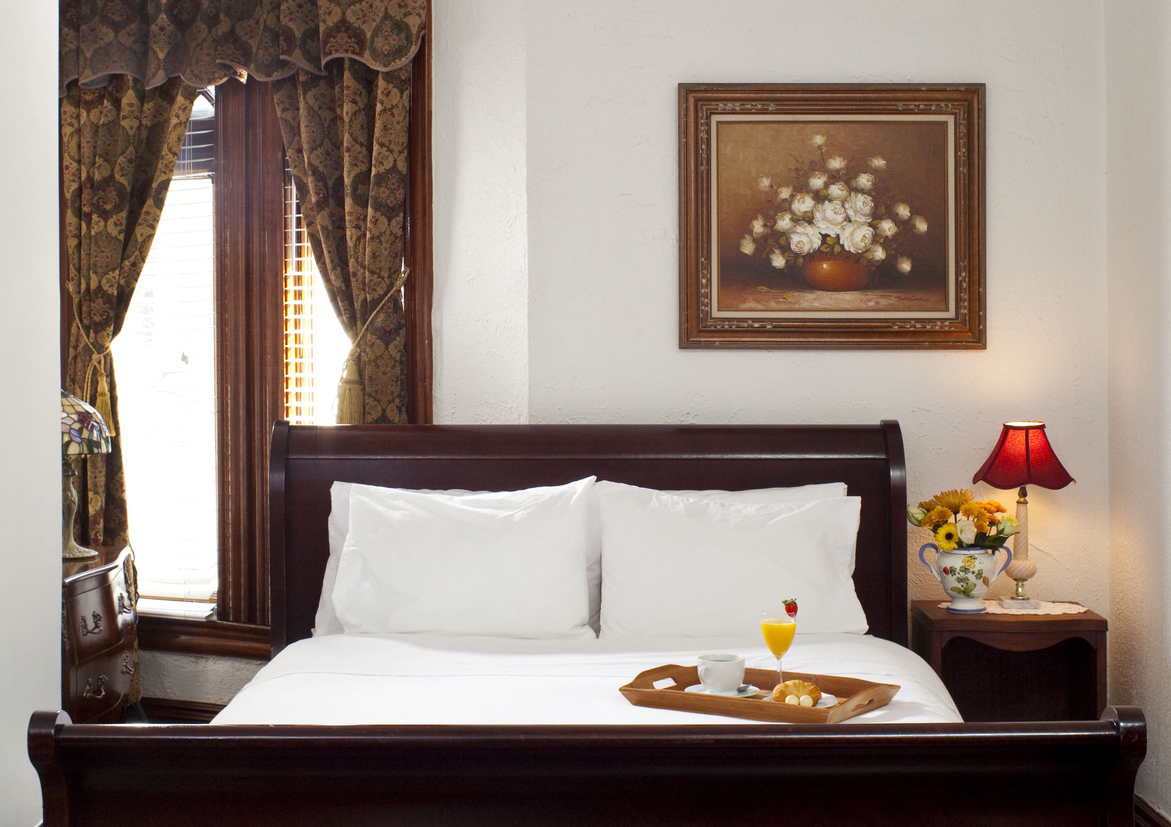 A neatly made bed with white linens, a tray with breakfast items, a painting of flowers on the wall, and a lamp on the nightstand.