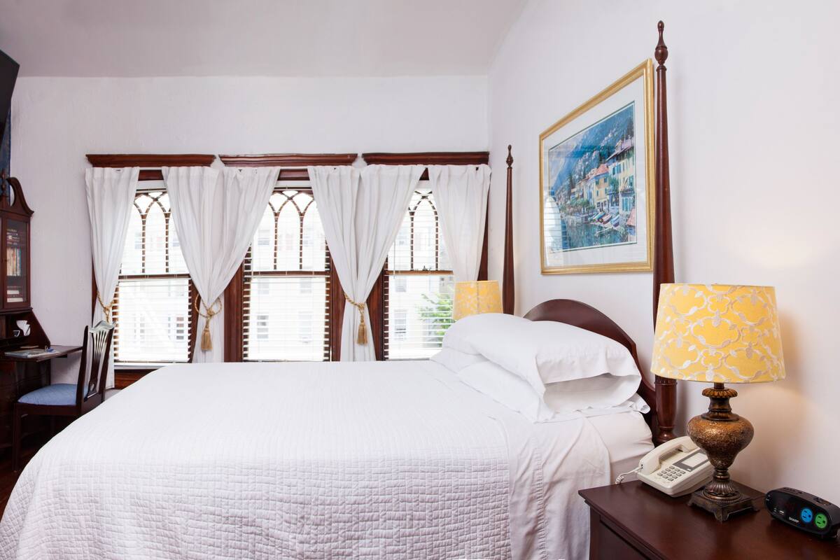 A bedroom with a large bed, a desk with a chair, a fireplace, and a window with curtains.