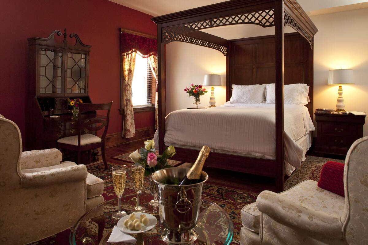 A four-poster bed with a nightstand and lamp, a dining table with chairs, and a cabinet.