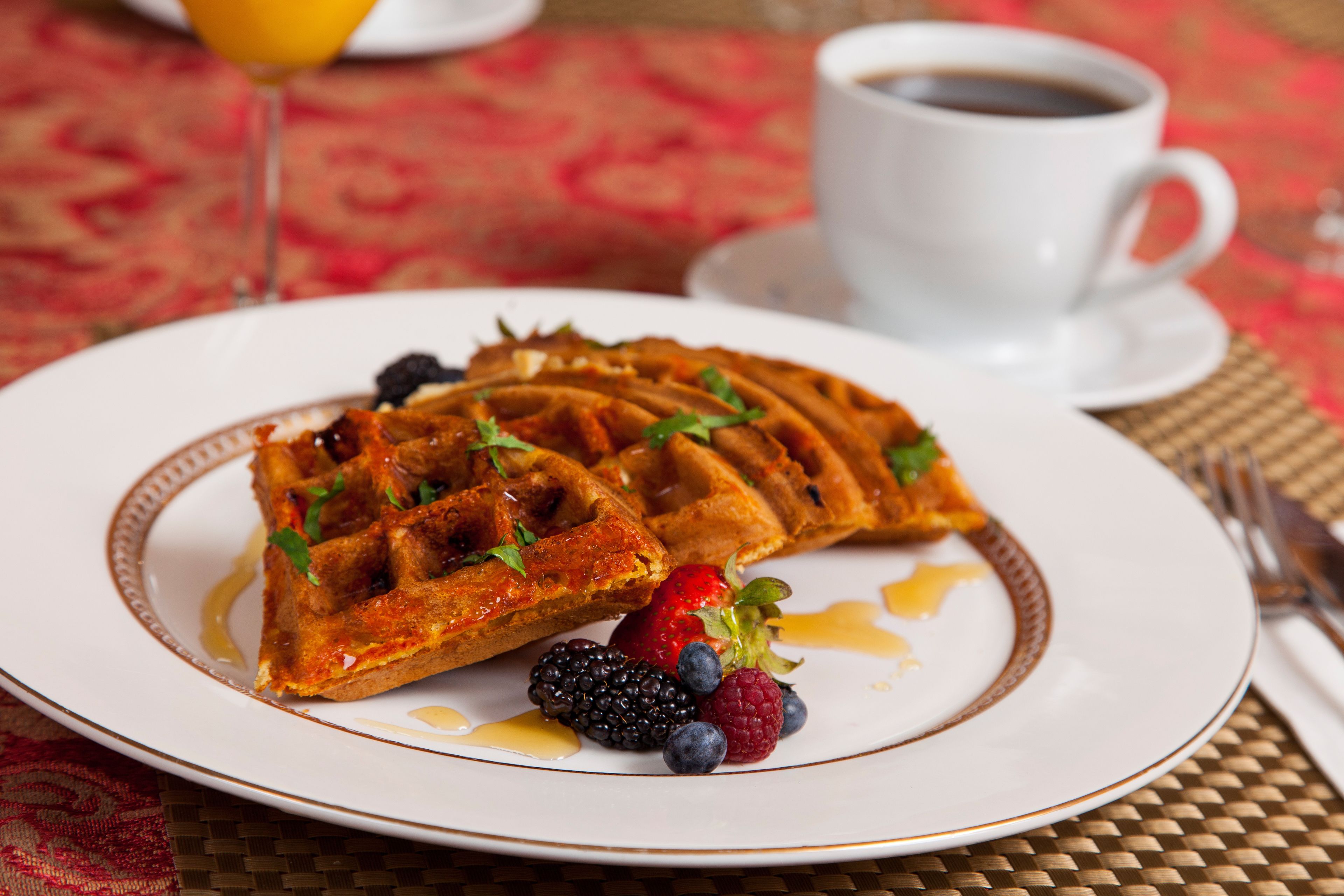 A plate of waffles with syrup and berries, a cup of coffee, and a glass of orange juice on a table with a red patterned tablecloth.