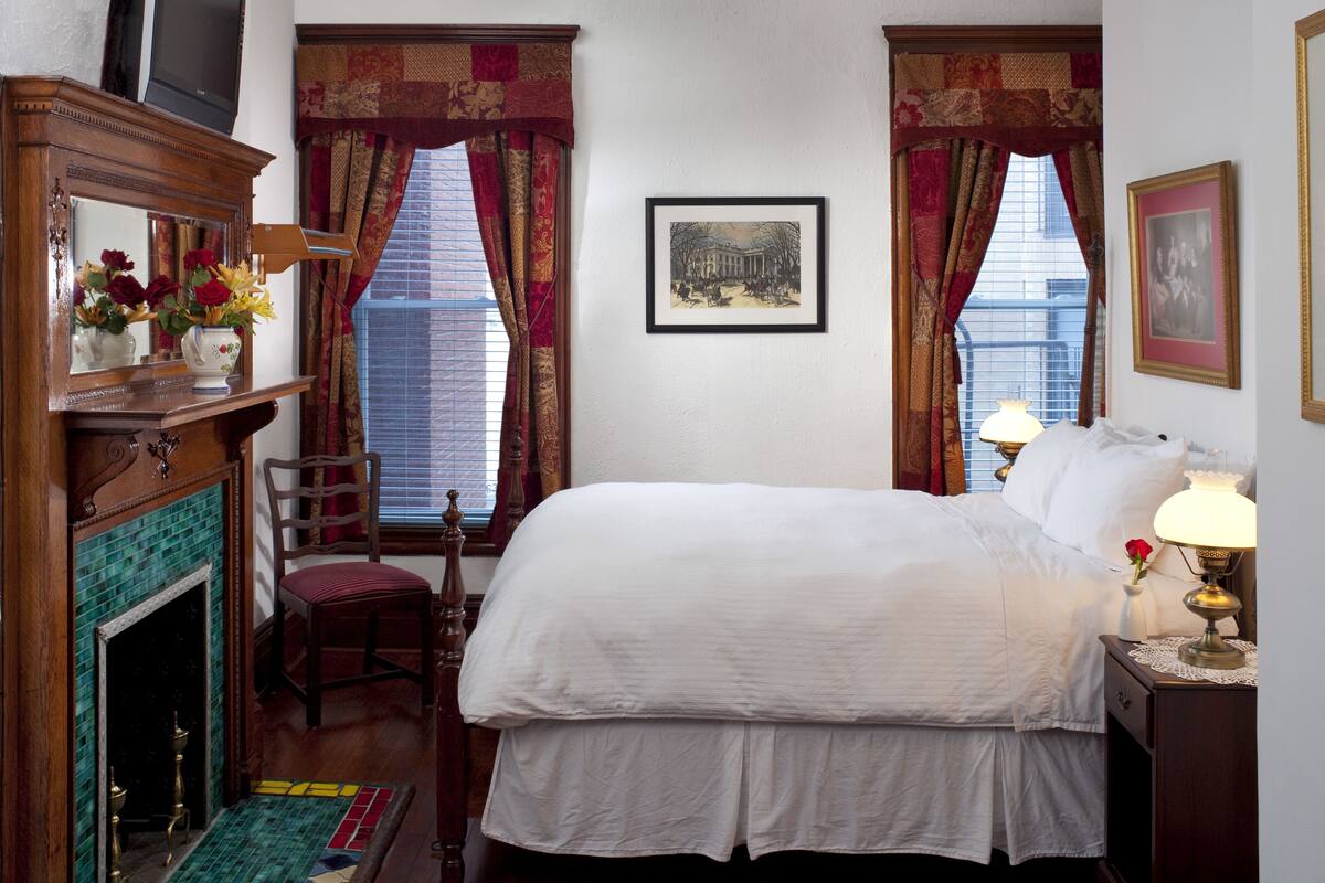 A hotel room with a bed, a chair, a fireplace, a picture on the wall, and a window with curtains.