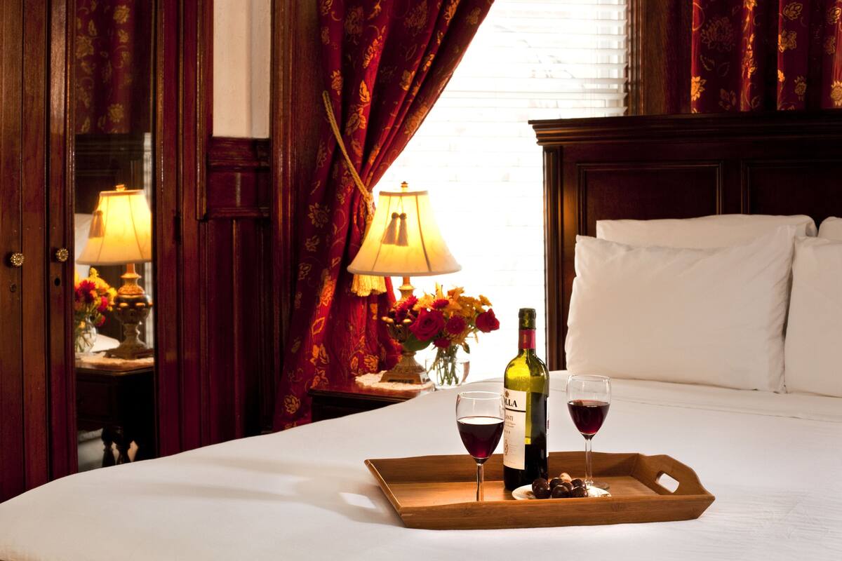 A hotel room with a bed, a bedside table with a lamp, a tray with a bottle of wine and a glass, and a vase with flowers.