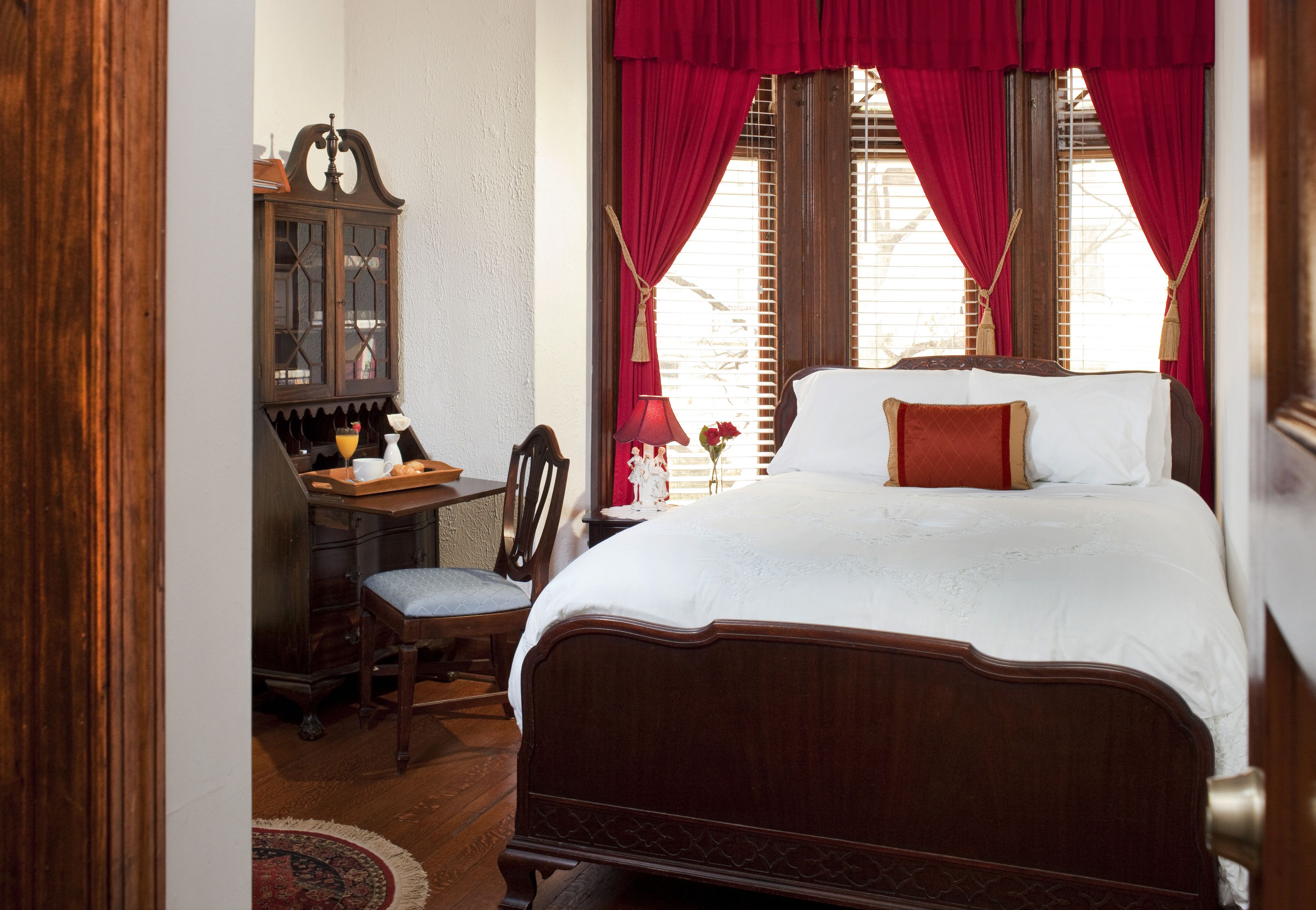 A bedroom with a wooden bed, red curtains, a wooden desk with a chair, and a small table with a vase of flowers.