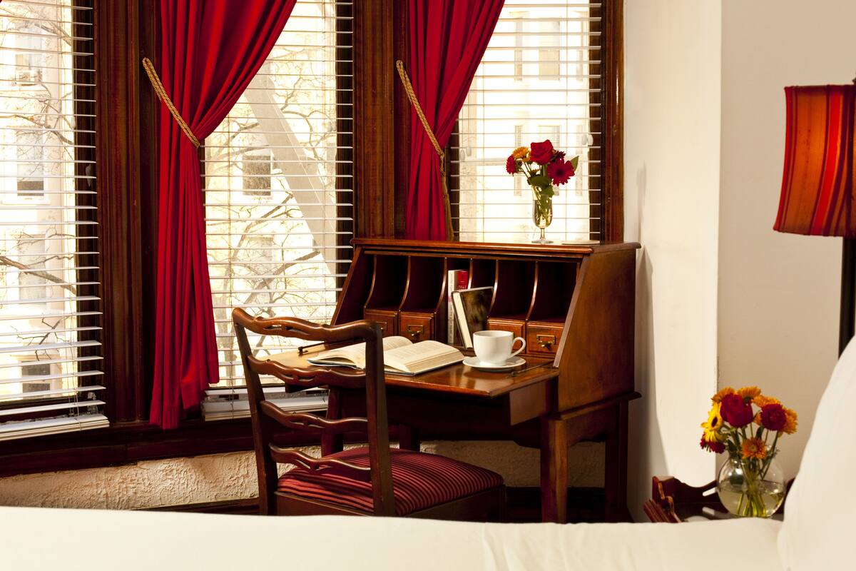 A hotel room with a wooden desk, a chair, a bed, a window with red curtains, and a vase with flowers.
