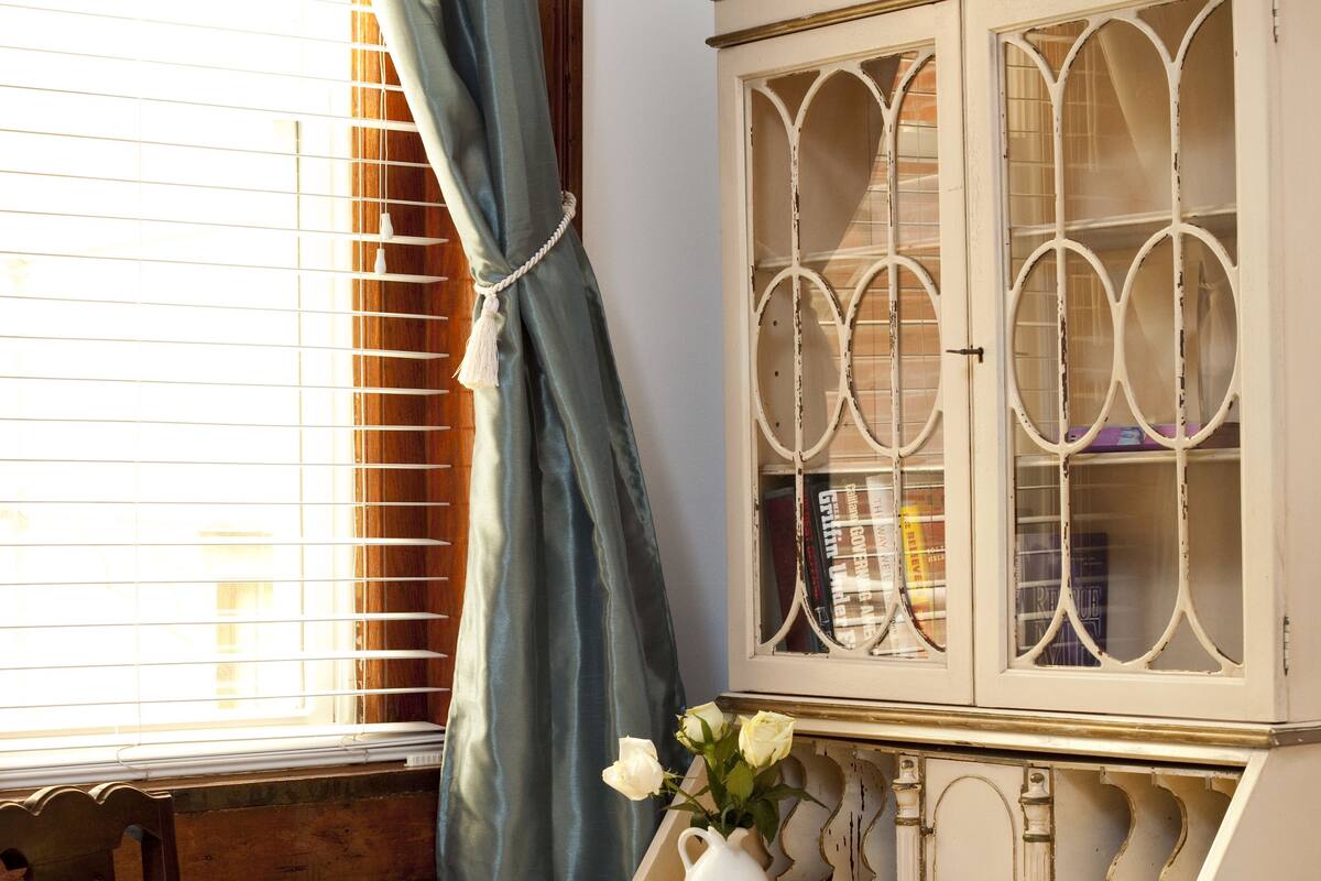 A vintage desk with a vase of white flowers, a chair with a patterned cushion, and a window with blinds.
