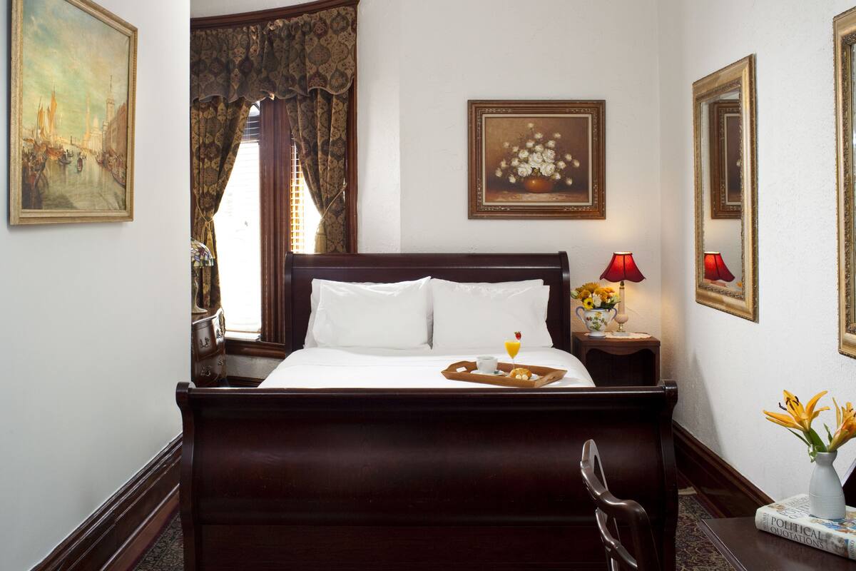A neatly made bed with white linens, a tray with a cup and a small dessert, a vase with flowers, and a framed painting on the wall.