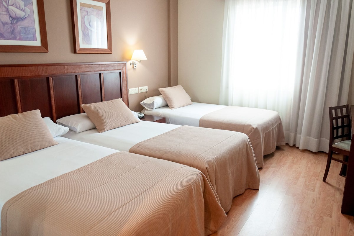 Habitación de hotel con dos camas, un cabecero de madera, una mesita de noche, una lámpara y una silla.