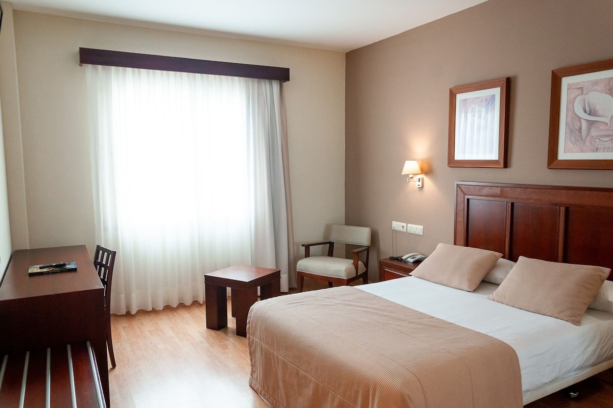 Habitación de hotel con cama, escritorio, silla, mesita de noche y lámpara.