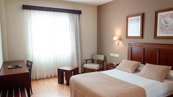 A hotel room with a bed, a desk, a chair, a nightstand, and a lamp.