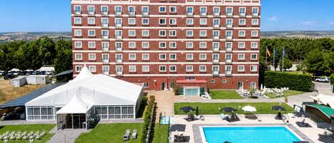 Un hotel de varios pisos con piscina, mobiliario exterior y una estructura tipo carpa blanca.