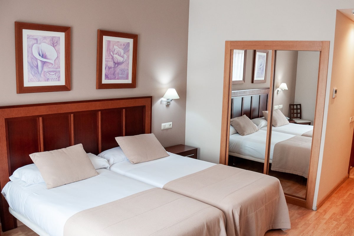 Una cama doble con cabecera de madera, dos mesitas de noche y una mesilla auxiliar. Se observan dos obras de arte enmarcadas en la pared sobre la cama.