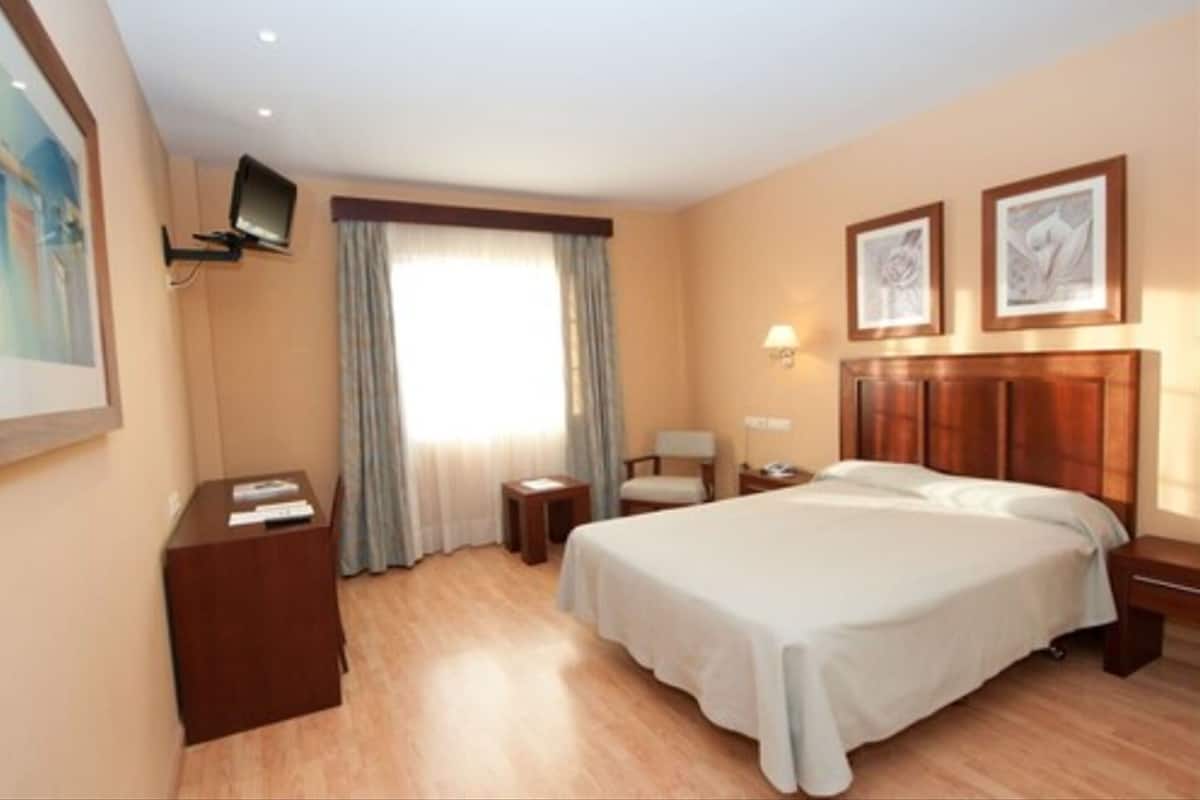 Habitación de hotel con cama, mesitas de noche, televisor montado en la pared y ventana con cortinas.