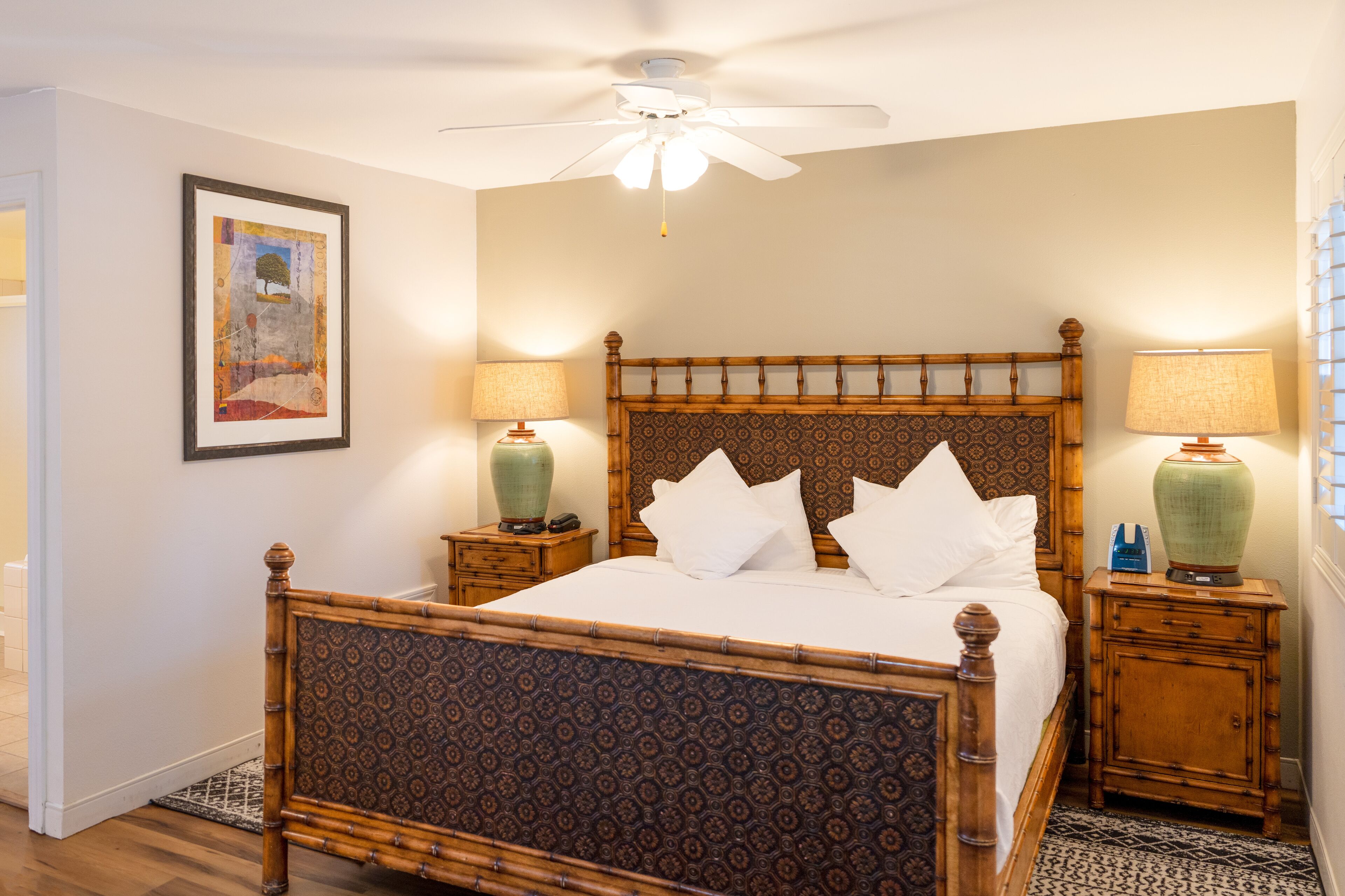 A neatly made bed with a wooden headboard, two bedside tables with lamps, a ceiling fan, and a framed picture on the wall.