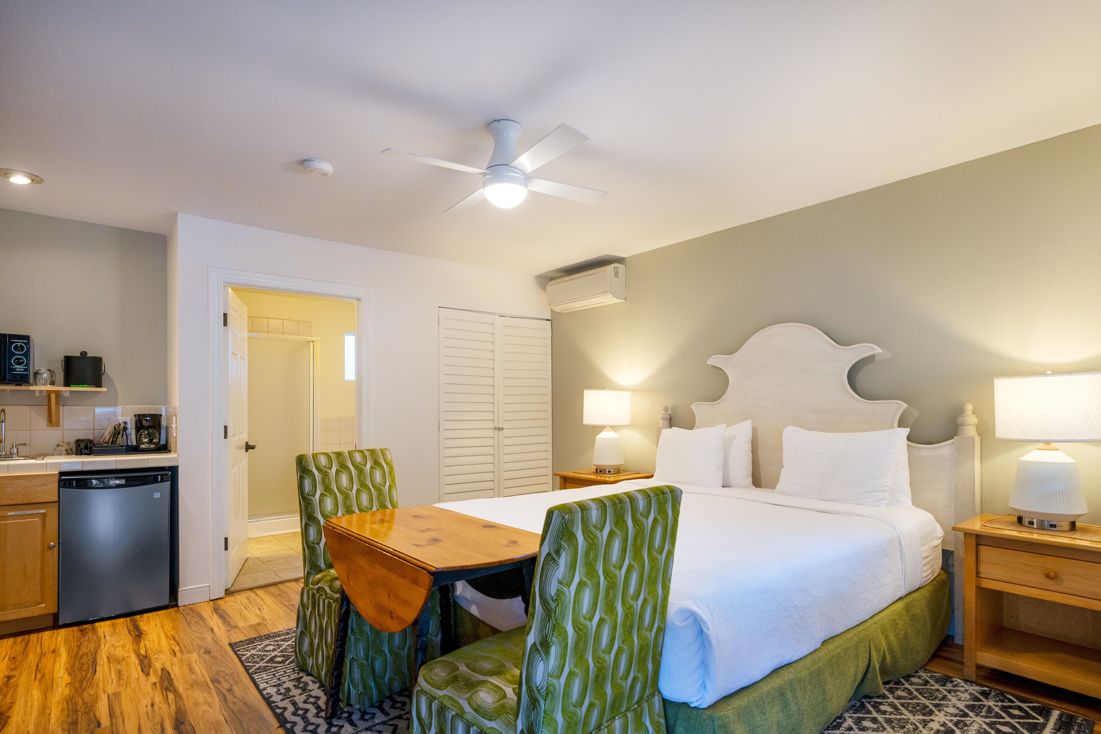 A hotel room with a bed, a desk, a chair, a ceiling fan, a bedside table, and a lamp.