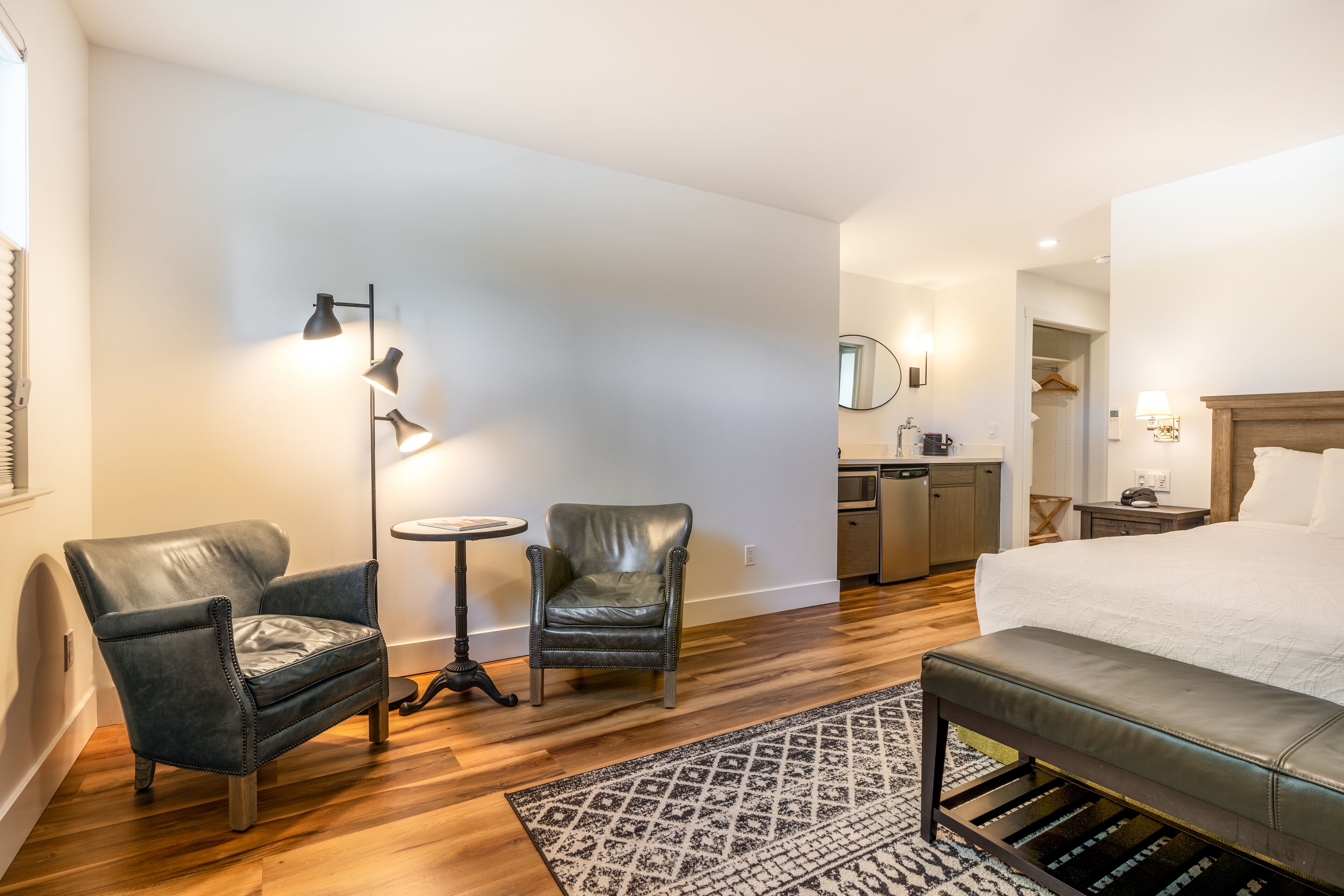 A hotel room with a bed, two armchairs, a small table, a floor lamp, a kitchen area, and a bathroom.
