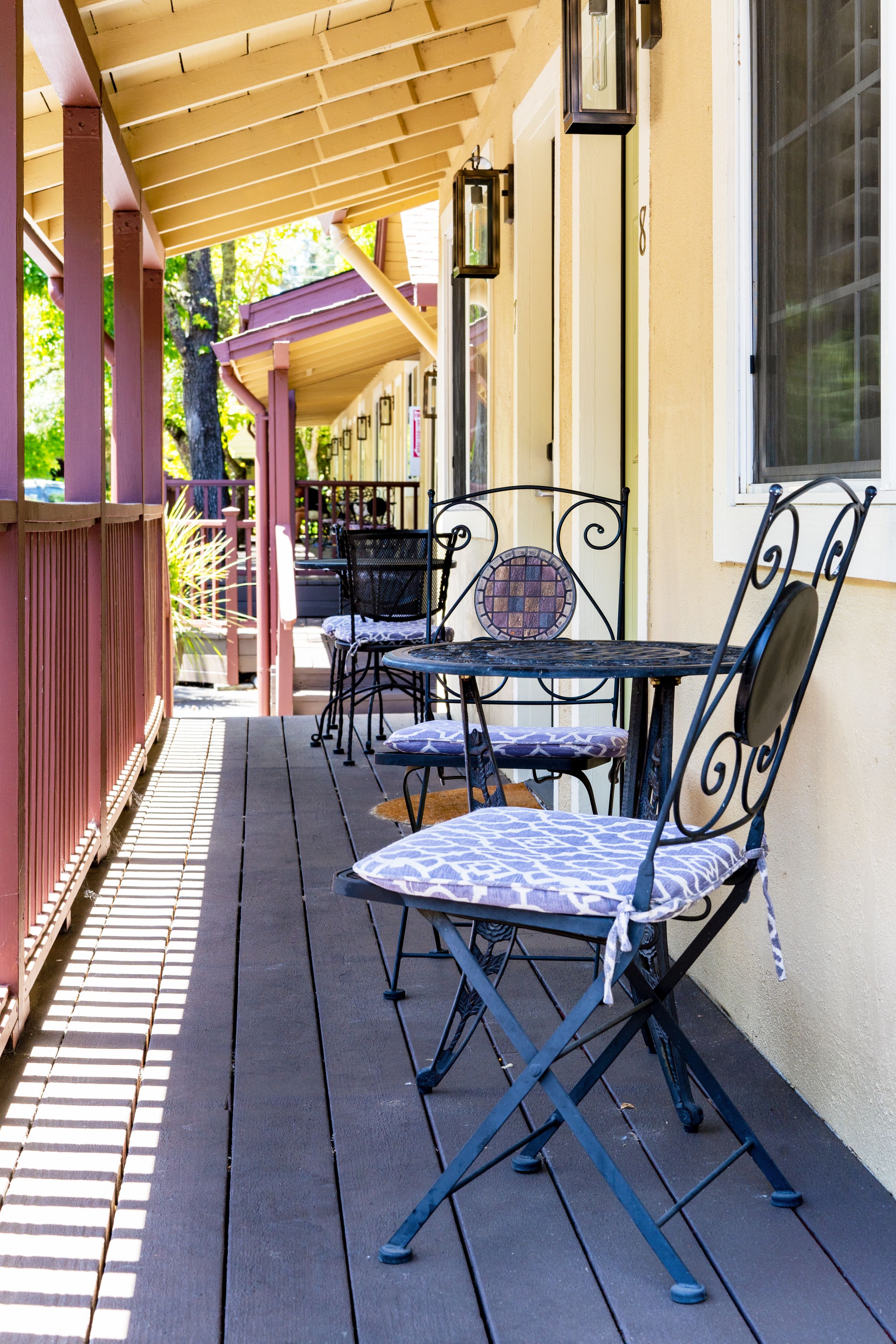 A covered outdoor seating area with metal chairs and tables, a wooden deck, and a building with a window and wall-mounted lights.
