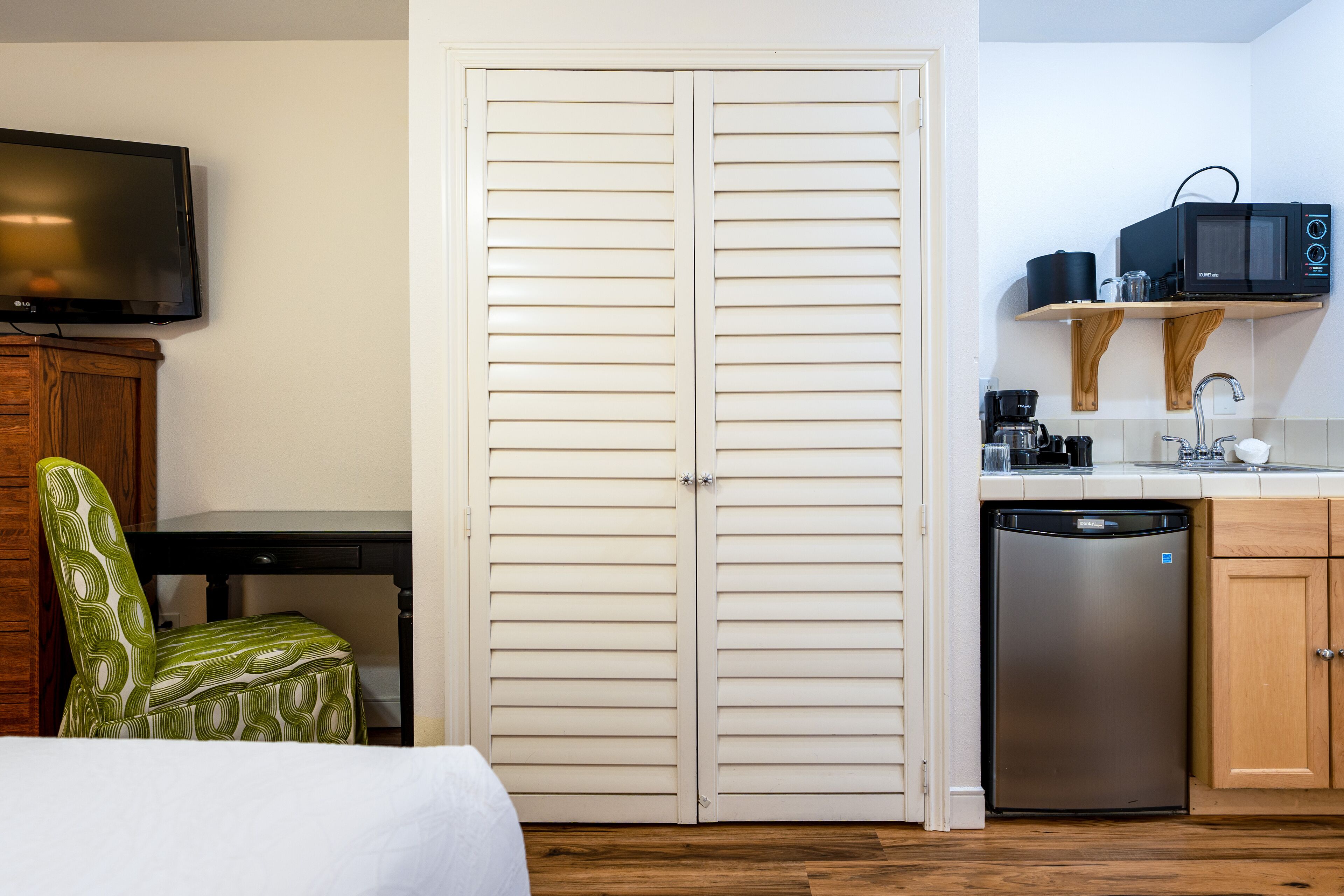 Habitación de hotel con cama, un sillón verde con estampado, un armario de madera, televisor, armario de doble puerta, y una zona de cocina con microondas, cafetera y lavavajillas.