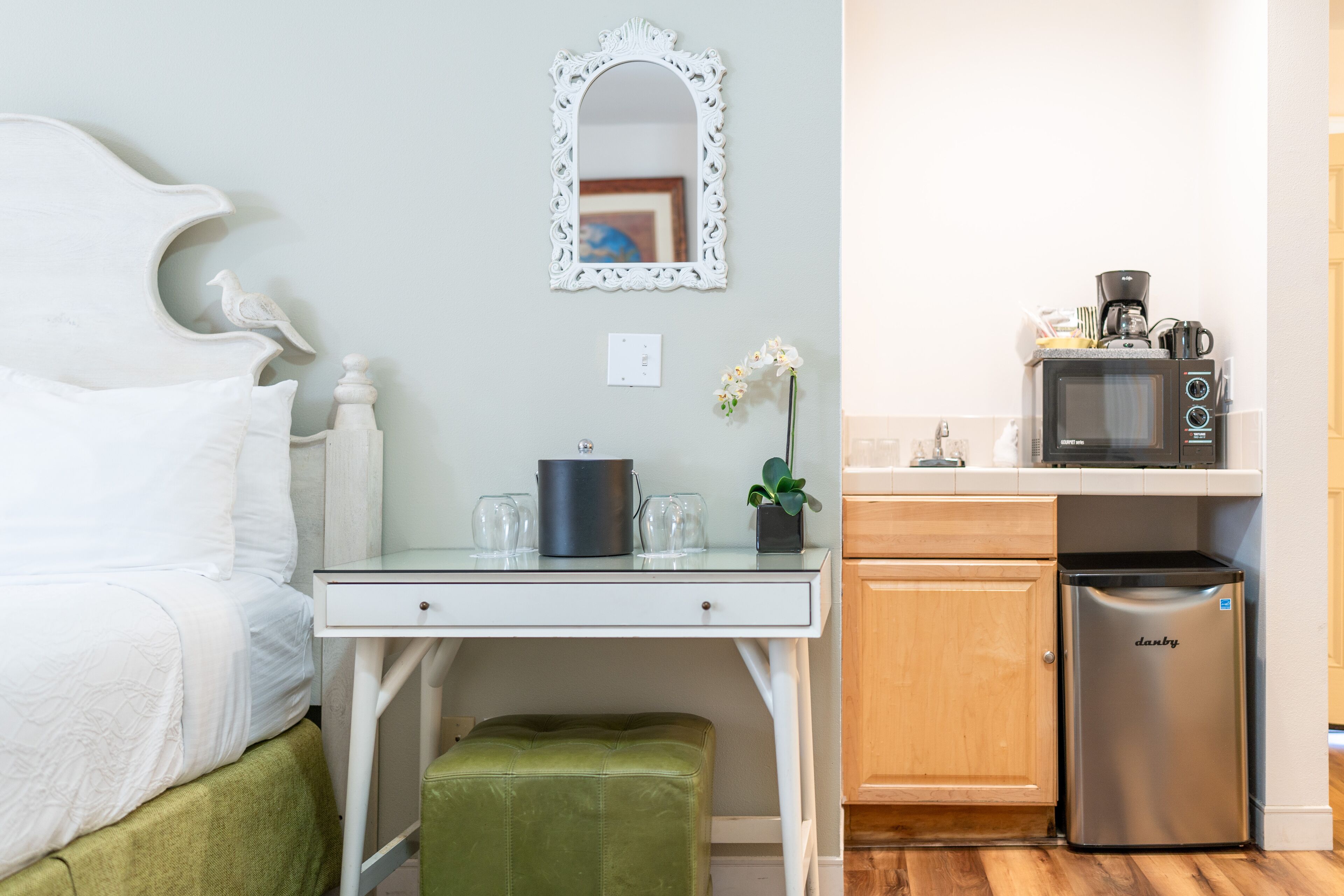 A bedroom with a bed, a small table with a lamp, and a green ottoman. The room has a white headboard and a decorative mirror on the wall.