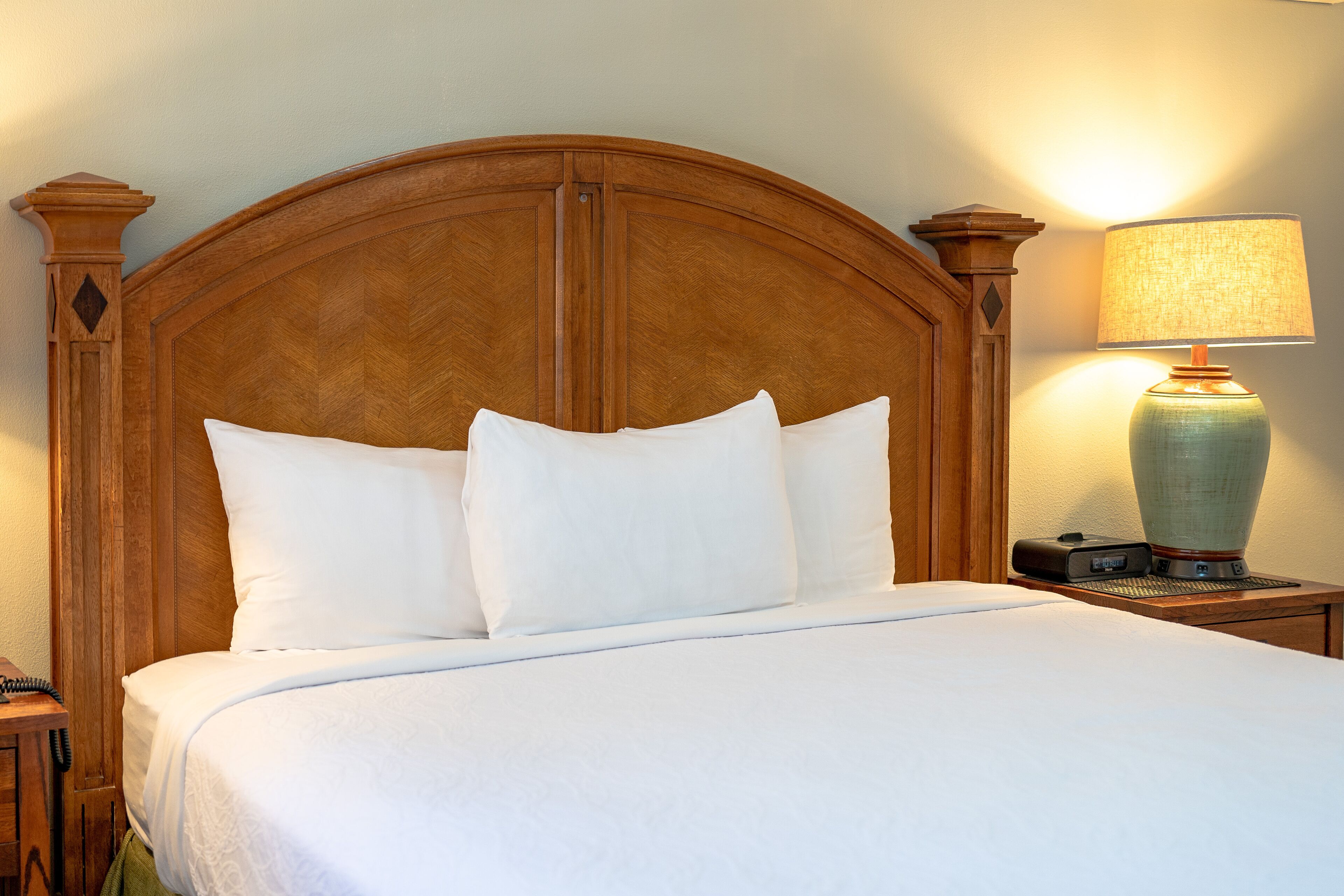 Een net opgemaakt bed met een houten hoofdbord, een bedlampje en een nachtkastje met een telefoon.