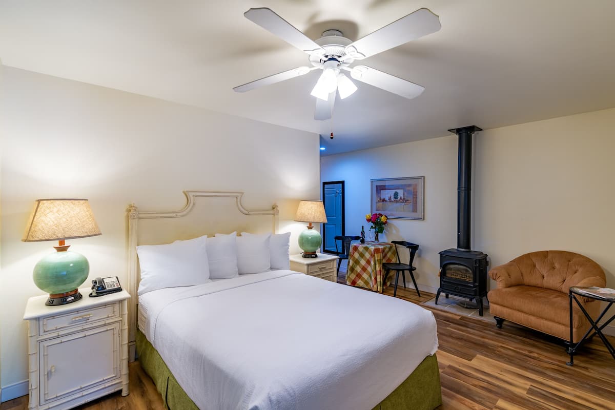 A neatly arranged bedroom with a bed, bedside lamps, a chair, a table, and a wood-burning stove.