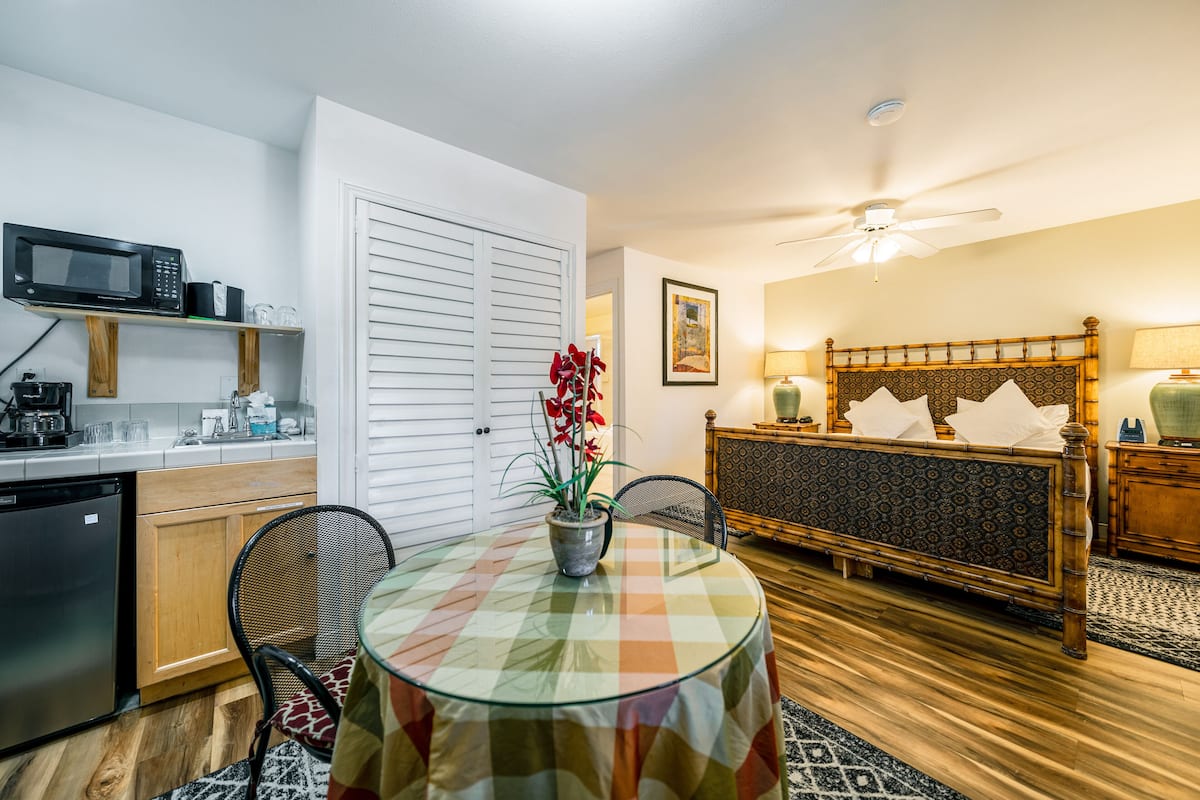 A compact room with a kitchen area, a dining table with a checkered tablecloth, a bed with a patterned headboard, and a ceiling fan.