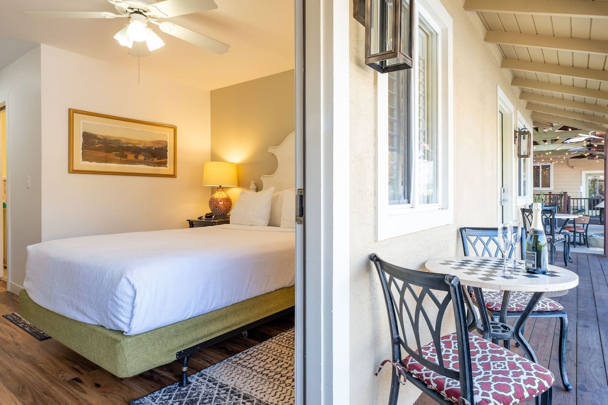 A hotel room with a bed, a nightstand, a lamp, and a view of the outdoor patio area with tables and chairs.