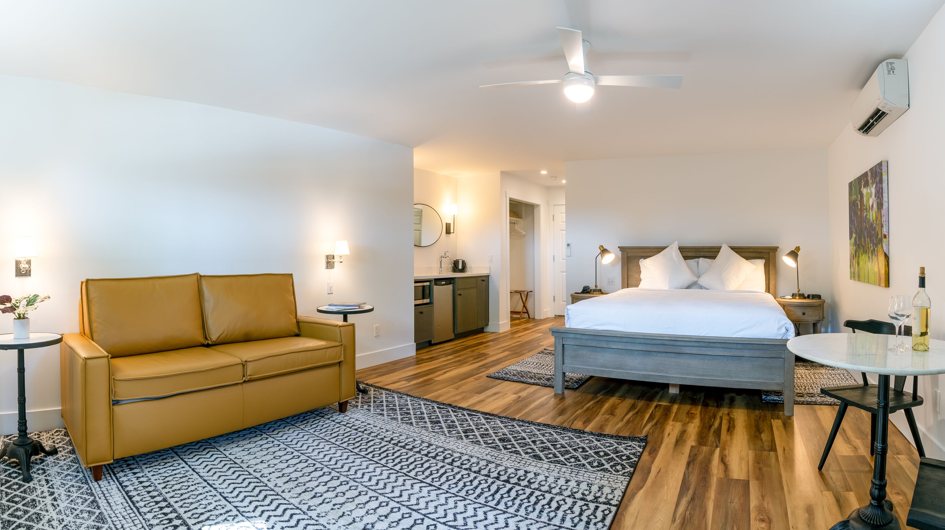 Una habitación de hotel moderna con una cama, un sofá, una mesita, un escritorio, un ventilador de techo, un equipo de aire acondicionado y un cuadro en la pared.