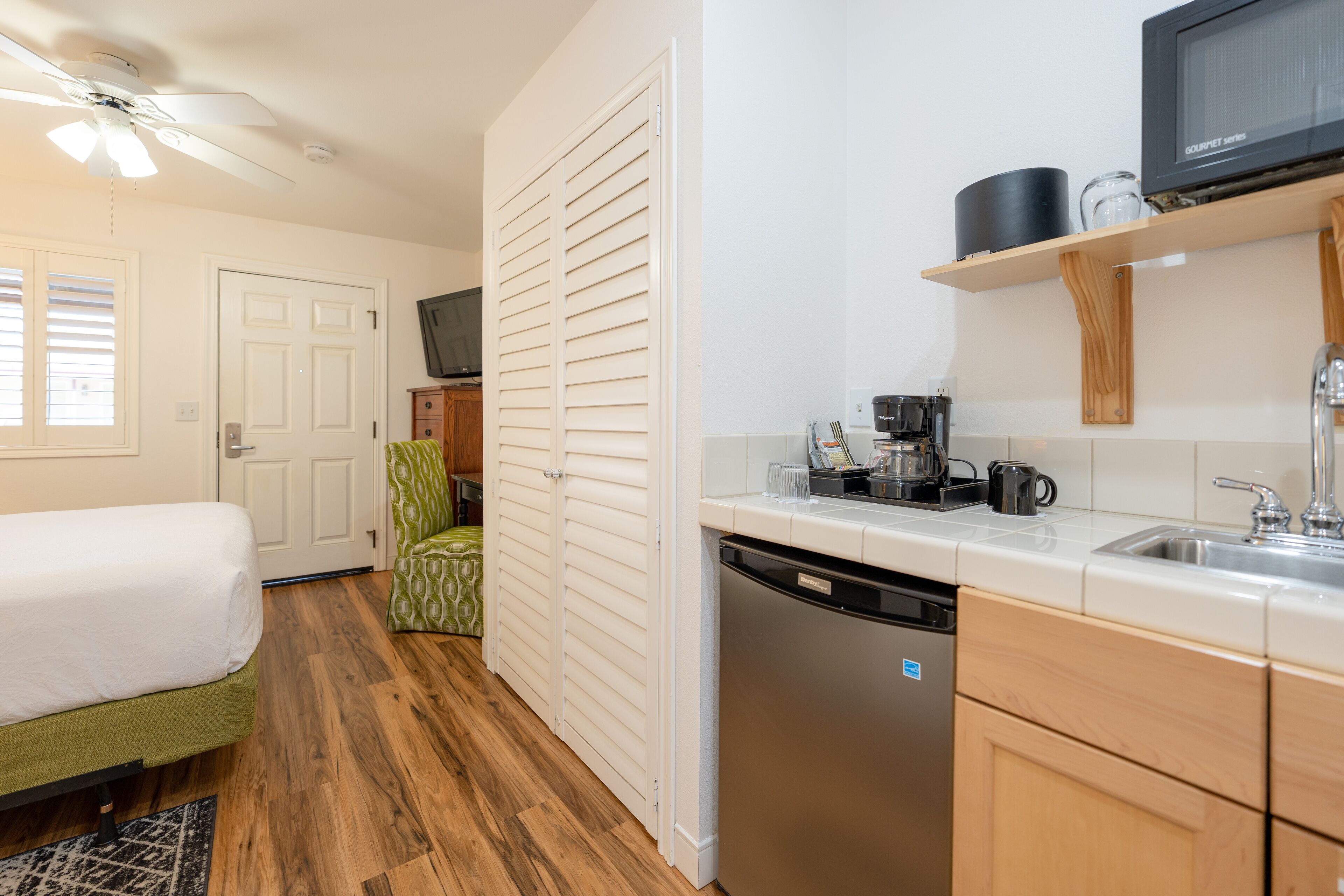 A compact hotel room with a bed, a green chair, a white wardrobe, a kitchen area with a dishwasher, and a ceiling fan.
