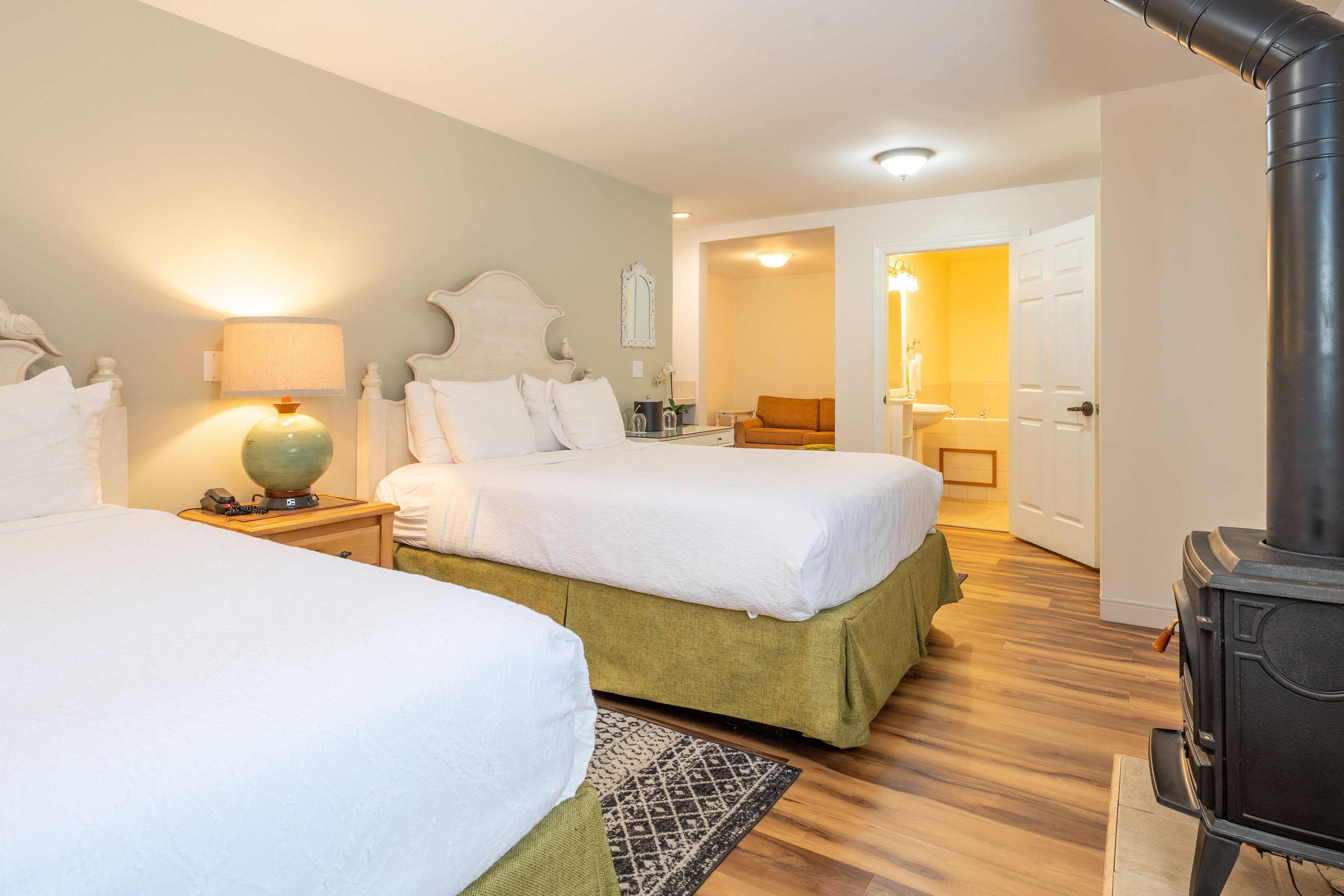 A hotel room with two beds, a wooden floor, a black stove, and a bathroom visible through an open door.