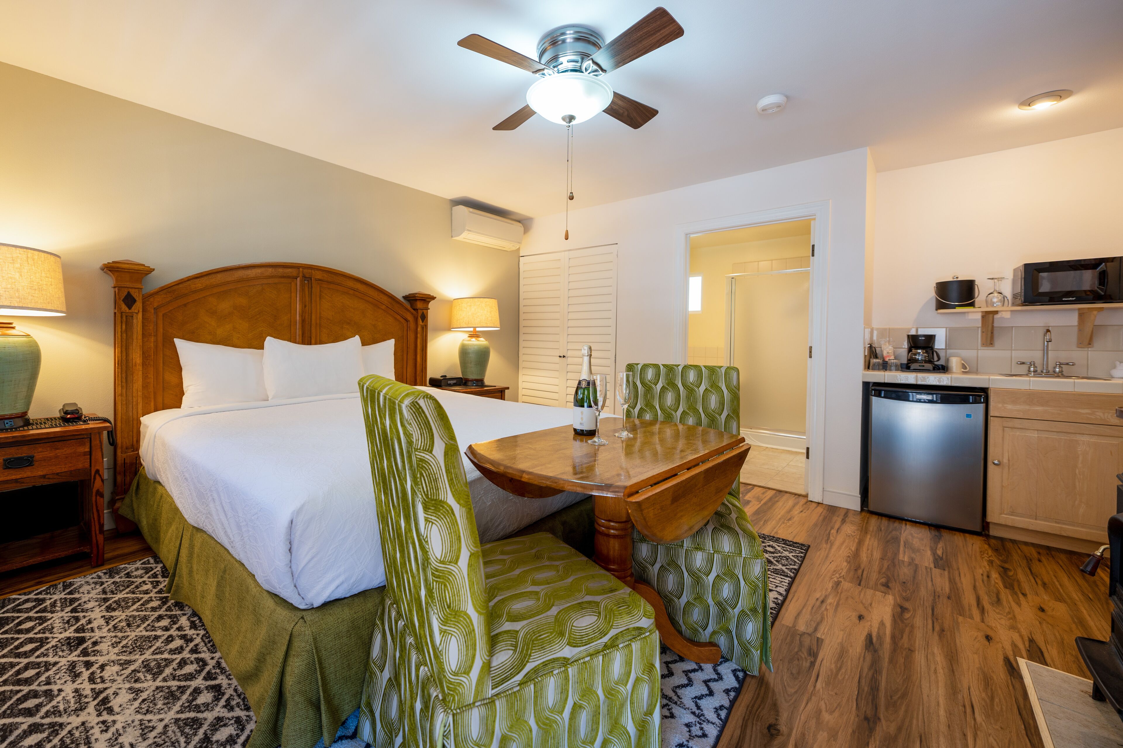 A hotel room with a bed, green patterned chairs, a wooden table, a ceiling fan, a kitchen area with a microwave, and a dishwasher.