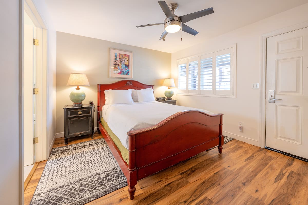 A bedroom with a wooden bed, two bedside tables with lamps, a ceiling fan, a window with blinds, and a door.