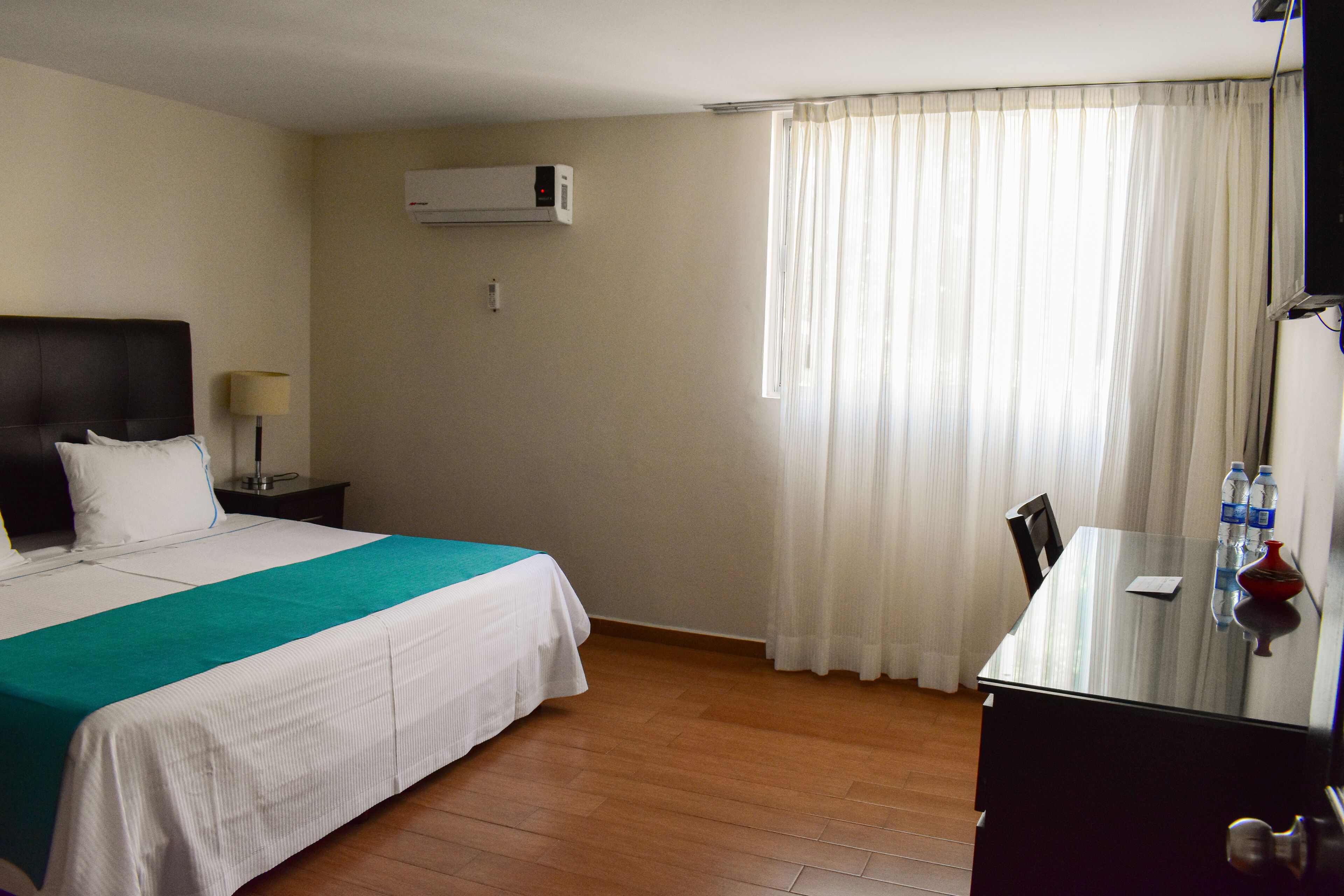 A hotel room with a bed, a desk, a chair, a lamp, and an air conditioning unit.